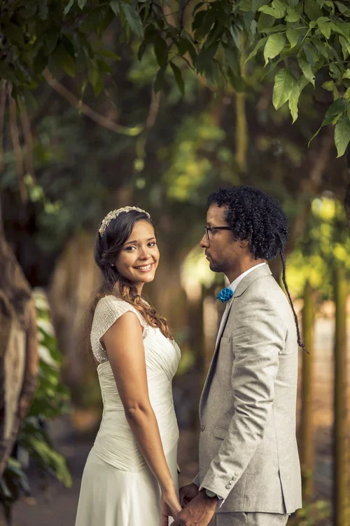 A couple dressed in wedding attire stands outdoors surrounded by trees, smiling and holding hands, with the woman looking at the camera and the man looking at her.