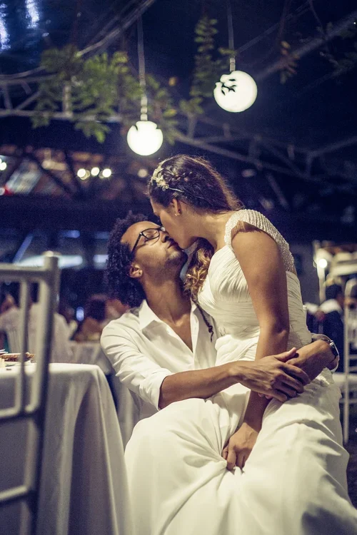 A couple sharing a romantic moment, with the woman sitting on a man's lap, both leaning in for a kiss, at an outdoor evening event with hanging lights.