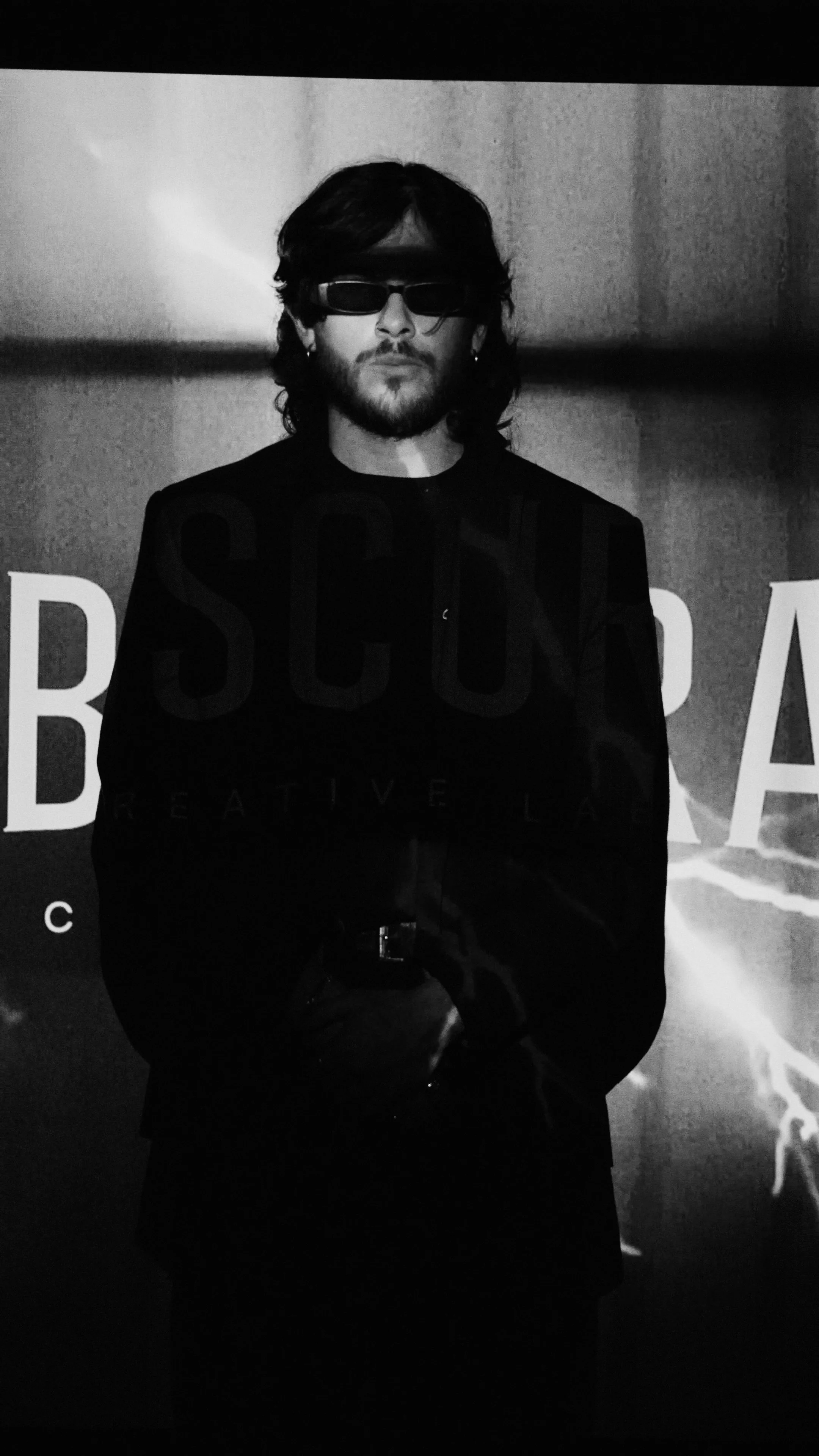 Black and white photo of a man with long hair, beard, and sunglasses, wearing a dark blazer and standing in front of a backdrop with large text.