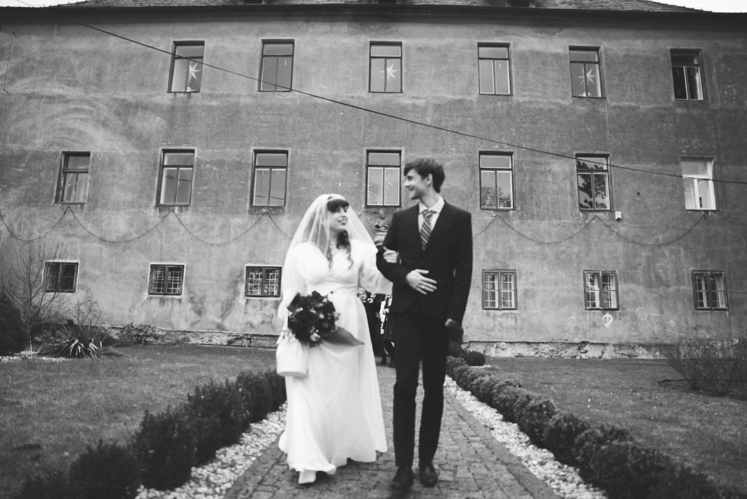 A black and white photo of a bride and groom walking outside on a garden path, with a large building in the background. The bride is wearing a wedding dress with a veil and holding a bouquet, while the groom is dressed in a suit and tie. They are smiling and looking at each other.