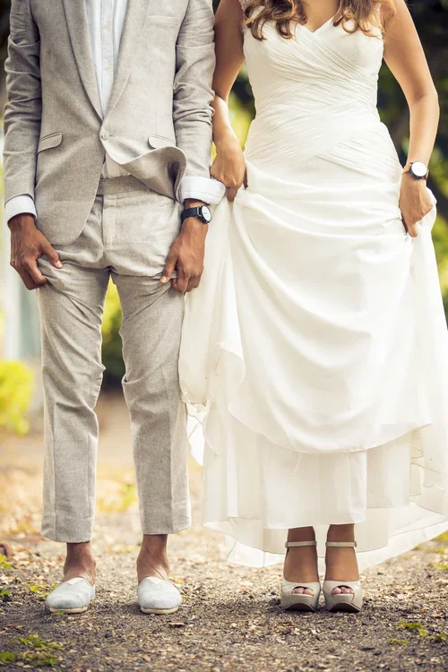 Couple holding hands, dressed in wedding attire, standing outdoors on a dirt path