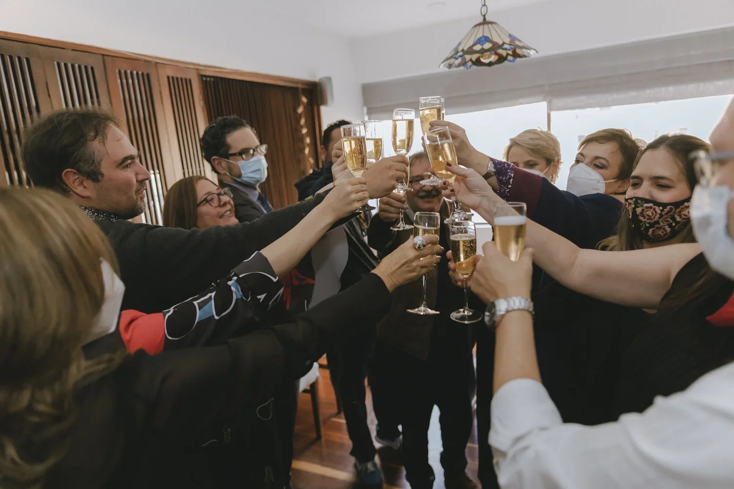 People wearing masks celebrating and raising glasses of champagne in a toast at a party indoors.
