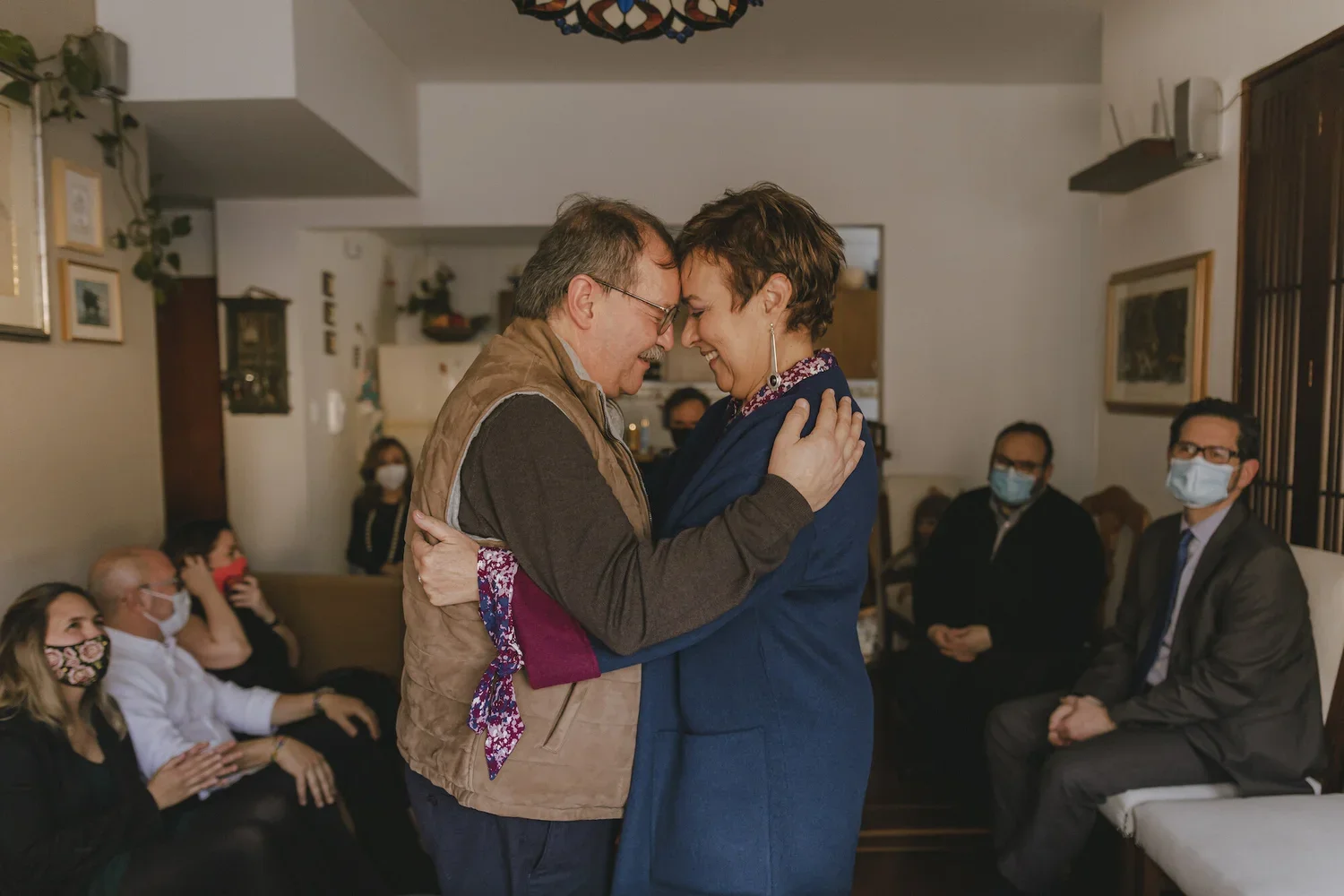 A man and woman hugging and smiling at each other in a room with seated people wearing masks in the background.