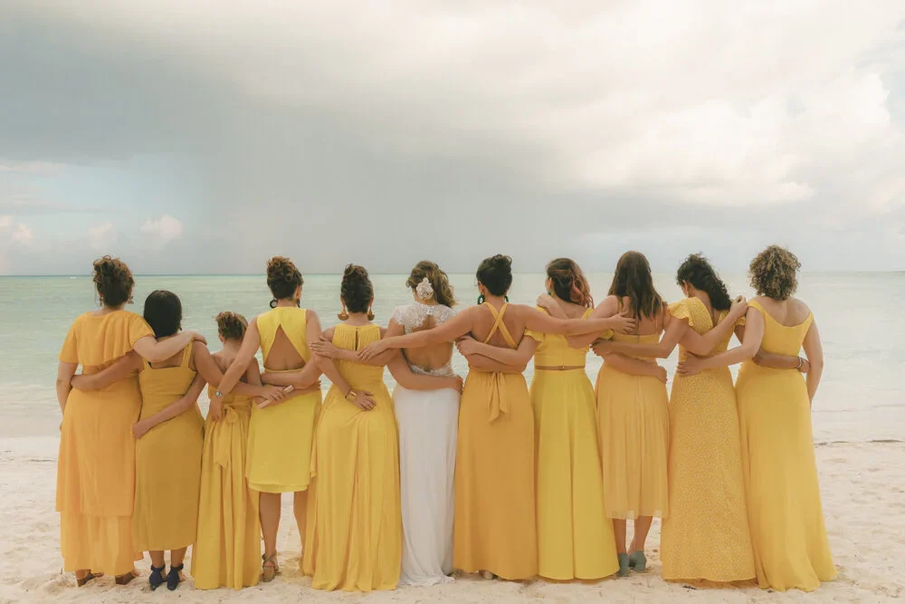 A group of eleven women in yellow dresses and one woman in a white dress, standing arm-in-arm on a beach, facing the ocean.
