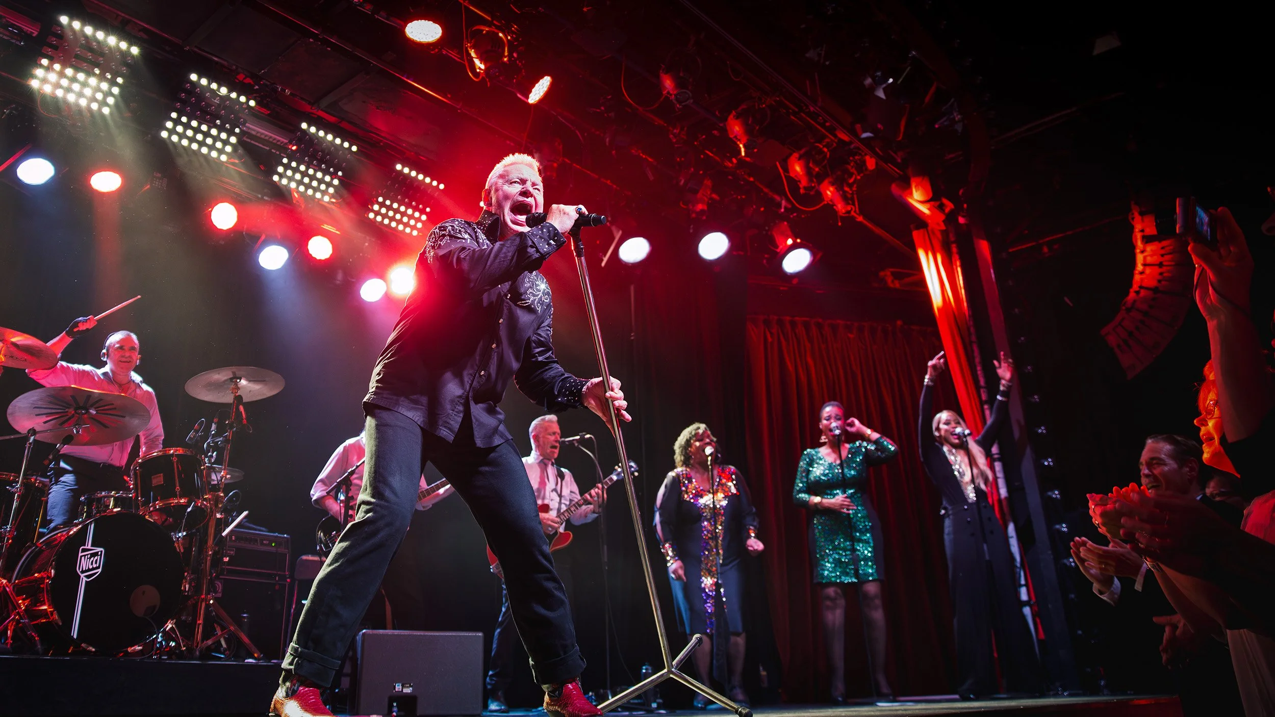 Jerry Williams holding a microphone stand and passionately singing, surrounded by backup singers and musicians under colorful stage lights in Stockholm 2016