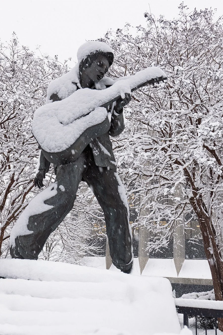 Statue of a person playing a guitar, covered in snow, outdoors with snow-covered trees in the background.