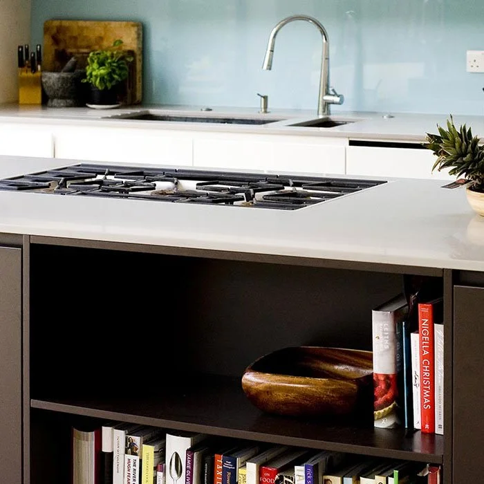 Modern kitchen with white countertops, a stovetop on an island, a sink with a silver faucet, green plant and cutting board in the background, and bookshelves with cookbooks and decorative items.