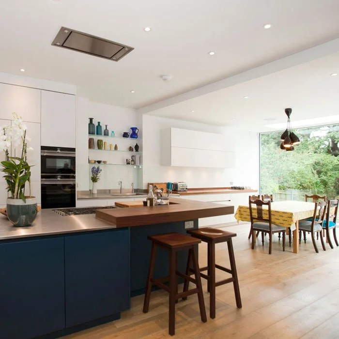Modern kitchen and dining area with blue lower cabinets, white upper cabinets, open shelves with colorful vases, wooden table with bench and chairs, large window overlooking greenery, and minimal lighting.