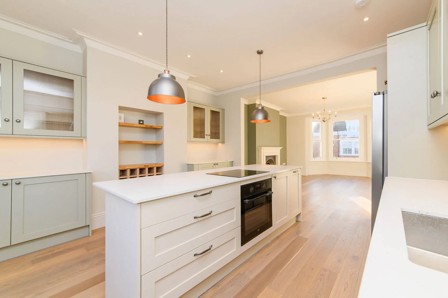 A spacious, modern kitchen with white cabinets, a central island, hardwood floors, and pendant lighting, opening into a living area with large windows and a fireplace.