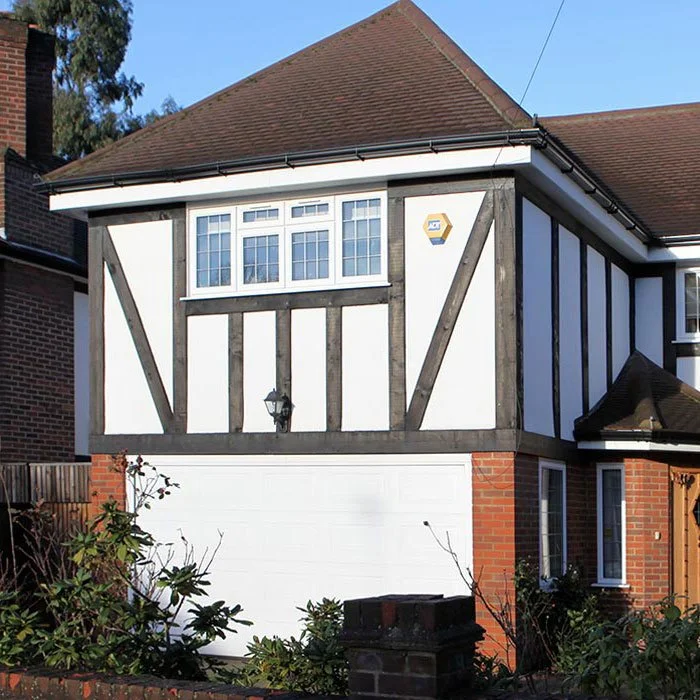 A two-story house with Tudor-style black and white framed woodwork, a red brick base, white garage door, and a small window with white trim on the second floor. There are plants in front and a lantern light beside the garage.
