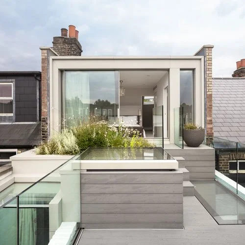 Modern rooftop terrace with glass railings, potted plants, and a view of neighboring buildings.