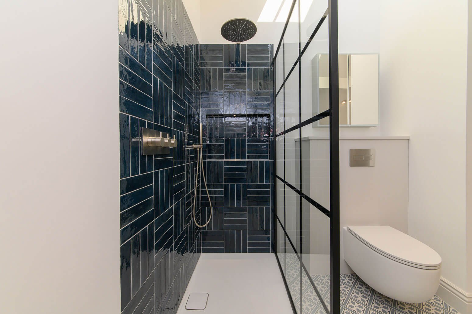 Modern bathroom with a walk-in shower featuring dark blue tiles arranged in various rectangular patterns, a ceiling-mounted rainfall shower head, and a handheld shower. To the right, a white toilet with a dual-flush system is positioned against the wall, which has a white cabinet with a mirror above it. The floor has patterned tiles in black, white, and gray. A glass partition with black framing separates the shower from the toilet.