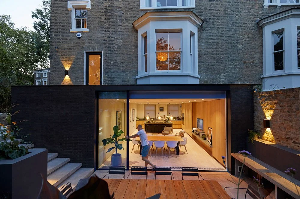 Person opening glass sliding doors to a modern backyard living space with dining area and TV
