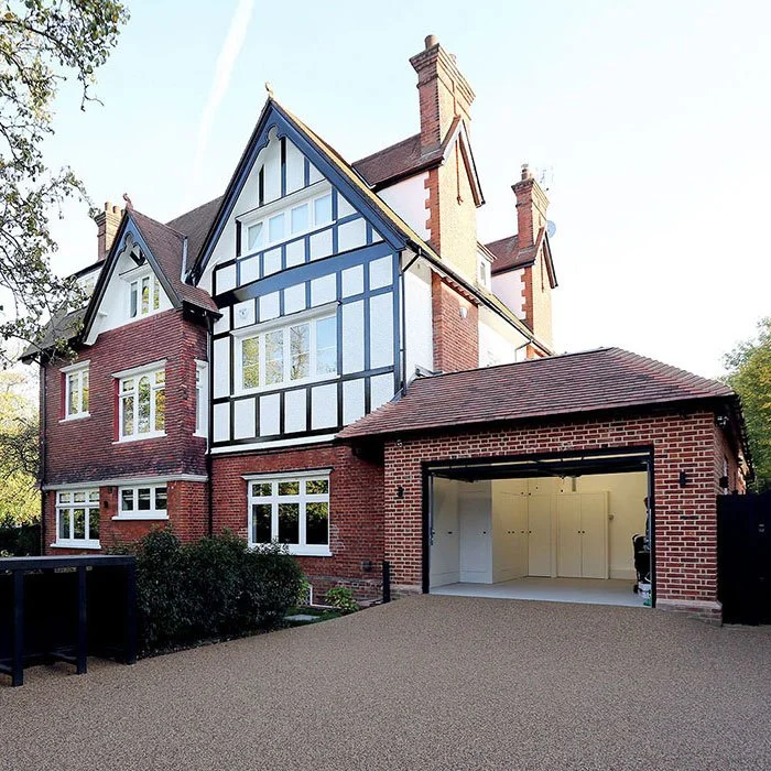 A large, multi-story brick house with Tudor-style design features, large windows, a garage with an open door, and a gravel driveway in a residential neighborhood.