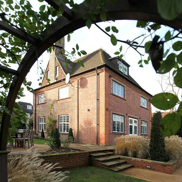 Brick house with multiple windows, a small front yard with bushes and grass, stone steps leading to the door, and surrounded by trees and plants.