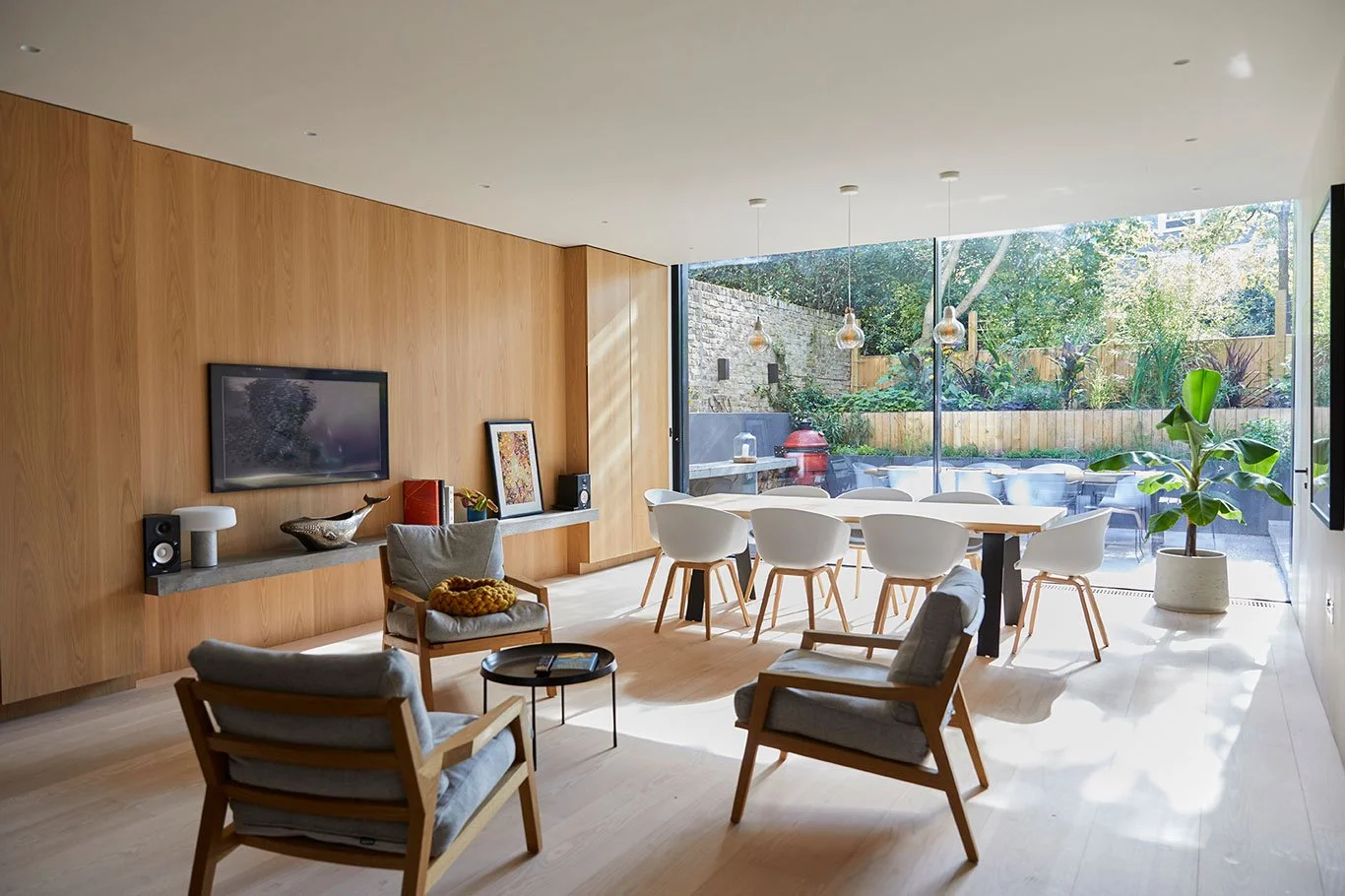 Modern living room with wooden wall, flat-screen TV, comfortable armchairs, coffee table, dining table with white chairs, large glass sliding doors opening to a garden with greenery, potted plant, and outdoor seating area.