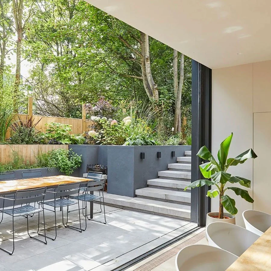 View of a modern indoor-outdoor living space with a sliding glass door opening to a backyard garden area with trees, plants, and a wooden fence. The interior features a table with chairs, a potted plant, and minimal decor.