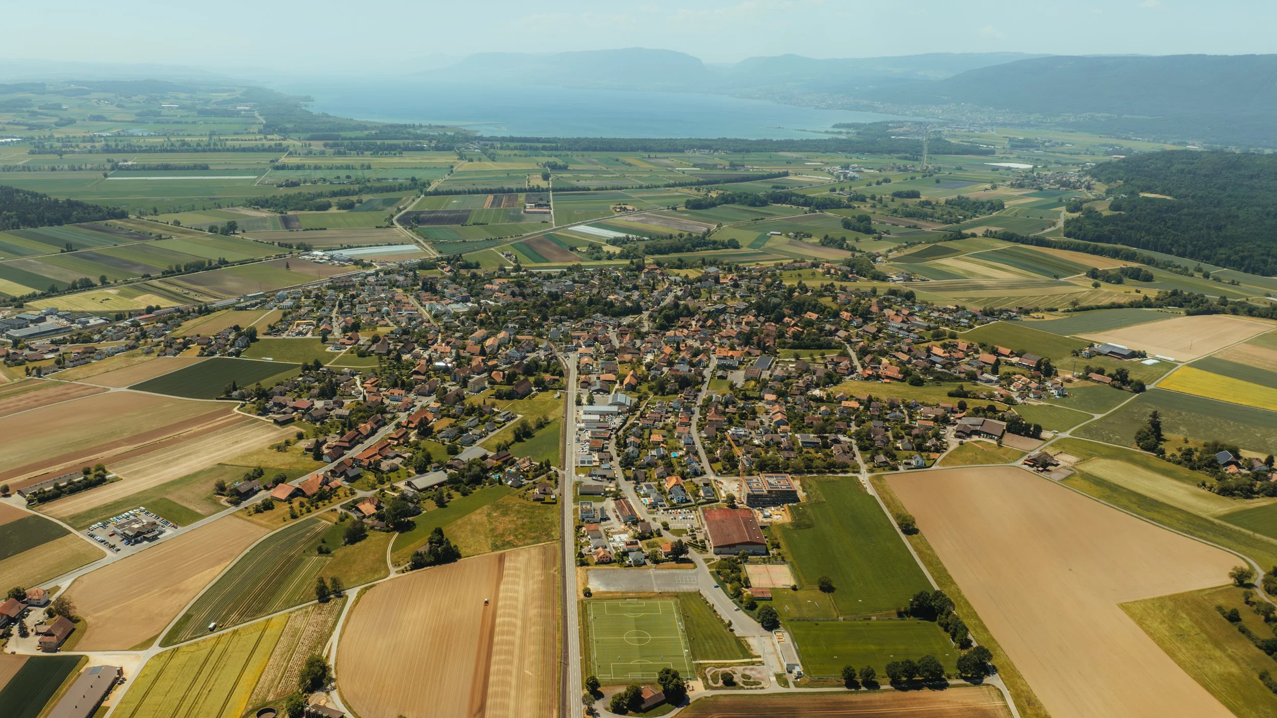 Luftaufnahme eines Dorfes mit landwirtschaftlichen Feldern, einem See im Hintergrund und umgeben von Hügeln.