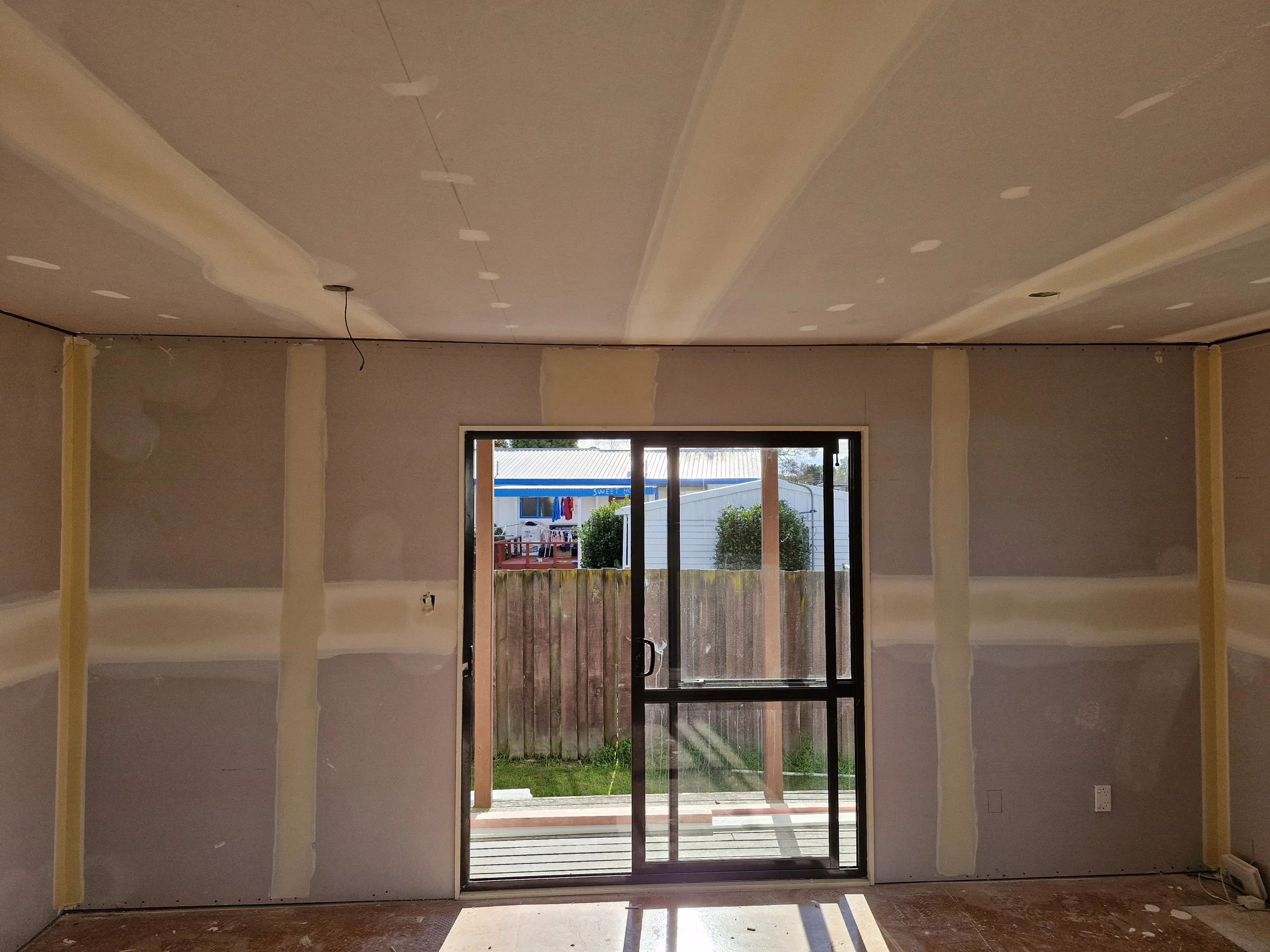 A room under construction with unfinished drywall, a ceiling with visible markings, and a sliding glass door leading to an outdoor area with a wooden deck and fence.