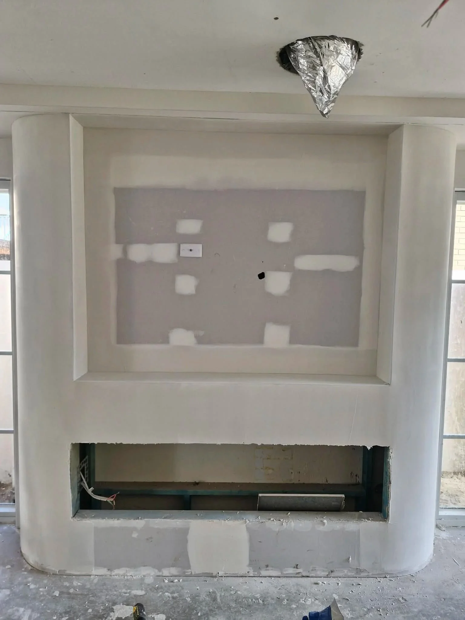 Fireplace structure under construction with a large rectangular opening at the bottom and a recessed area above, surrounded by white plaster walls, with unfinished surfaces and visible wiring inside the lower opening.