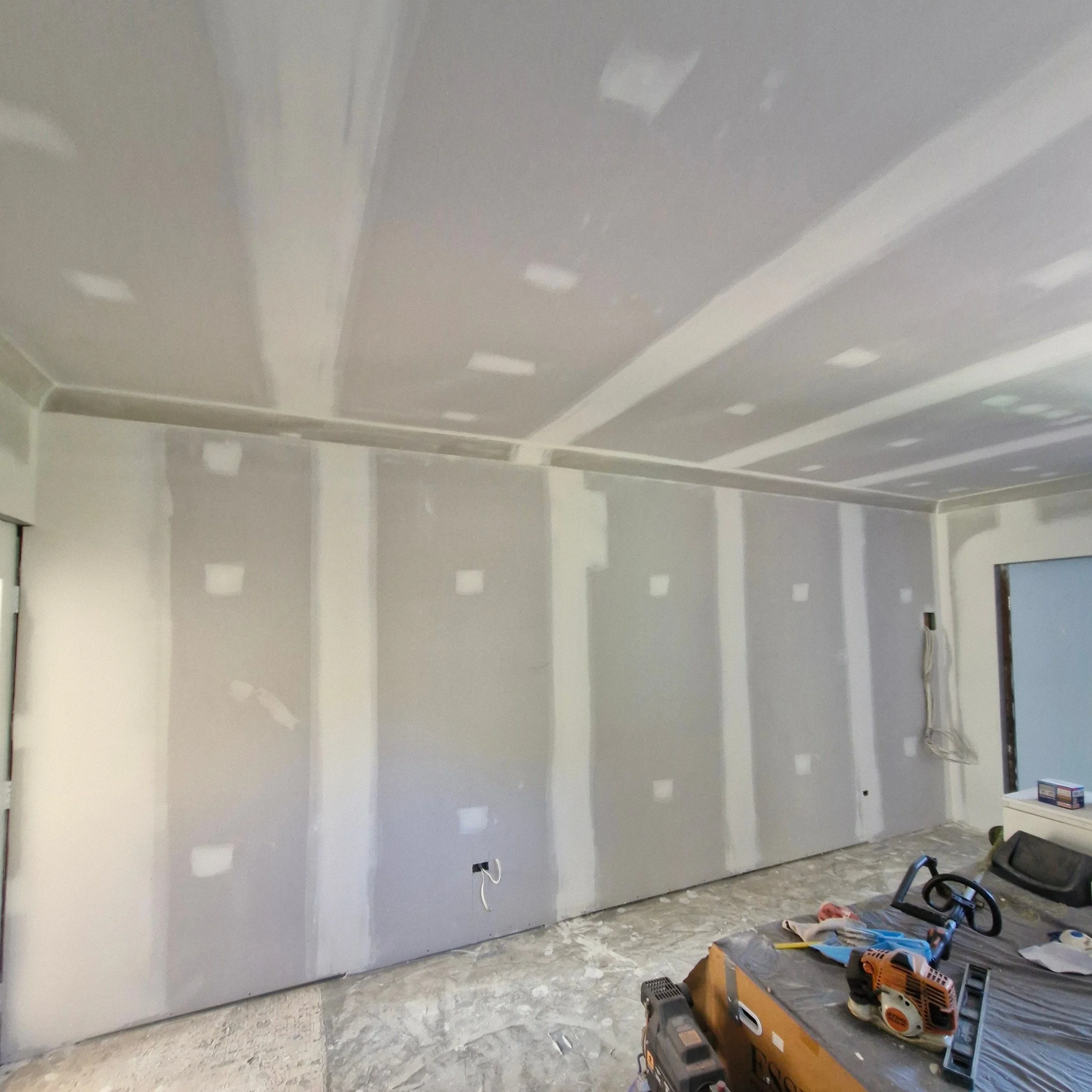 A room under construction or renovation with a white drywall wall and ceiling, unfinished floor, and various tools and equipment scattered around. The wall has visible seams and spots for future electrical outlets or switches.