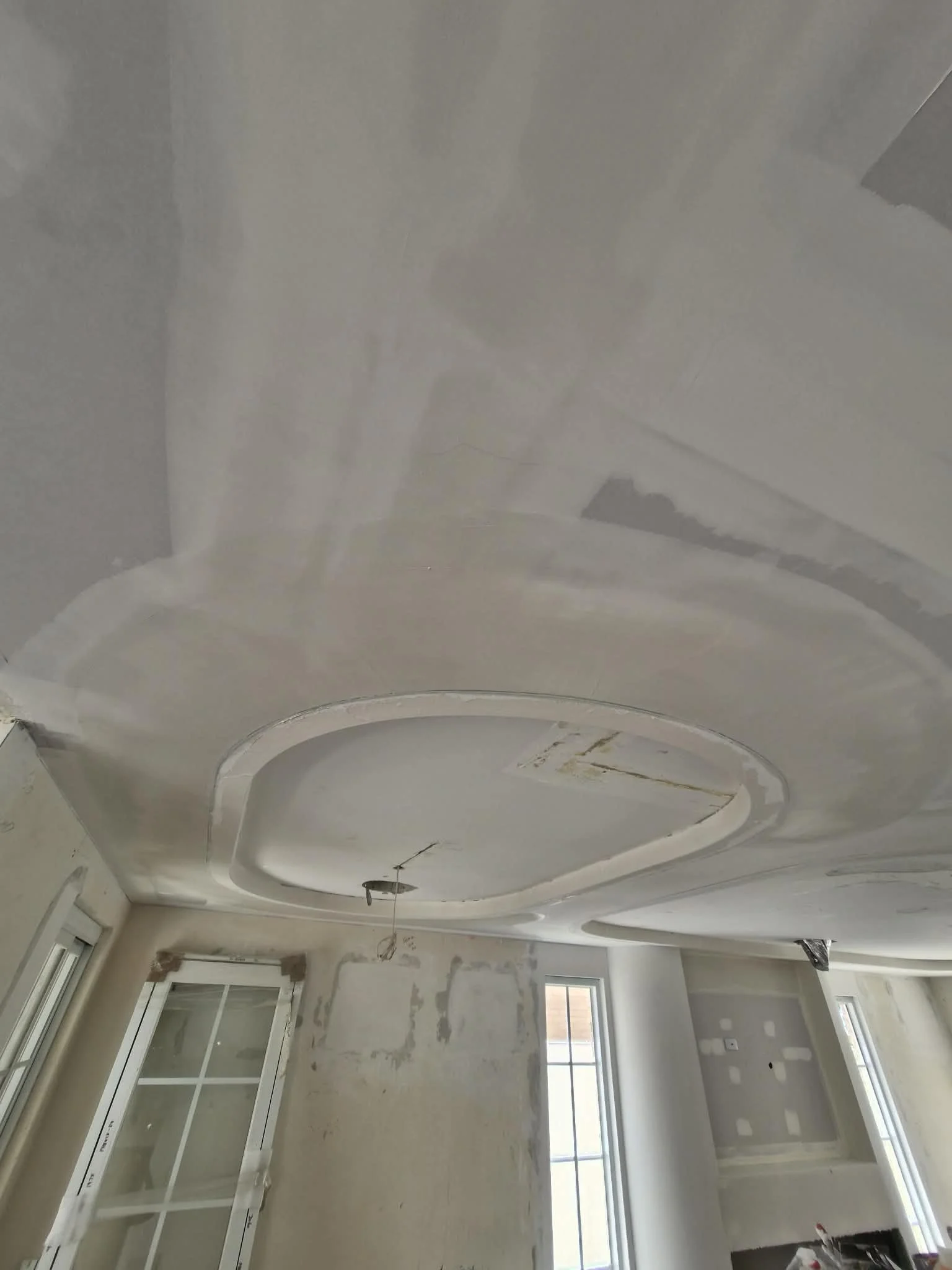 Interior of a room under construction with a newly installed ceiling featuring decorative molding, unfinished walls, and windows.
