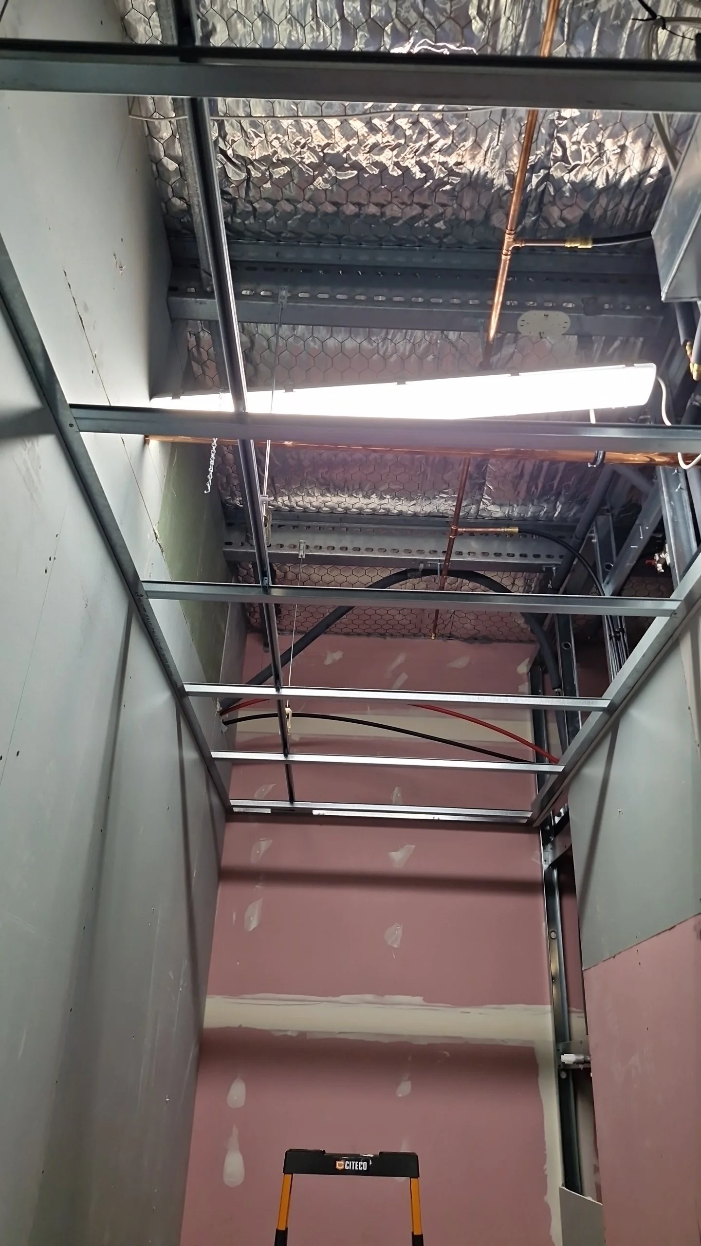 View of a construction site showing metal framing for a ceiling or wall, with electrical wiring and copper pipes installed. Part of the floor is visible with drywall patches, and a step ladder is in the foreground.
