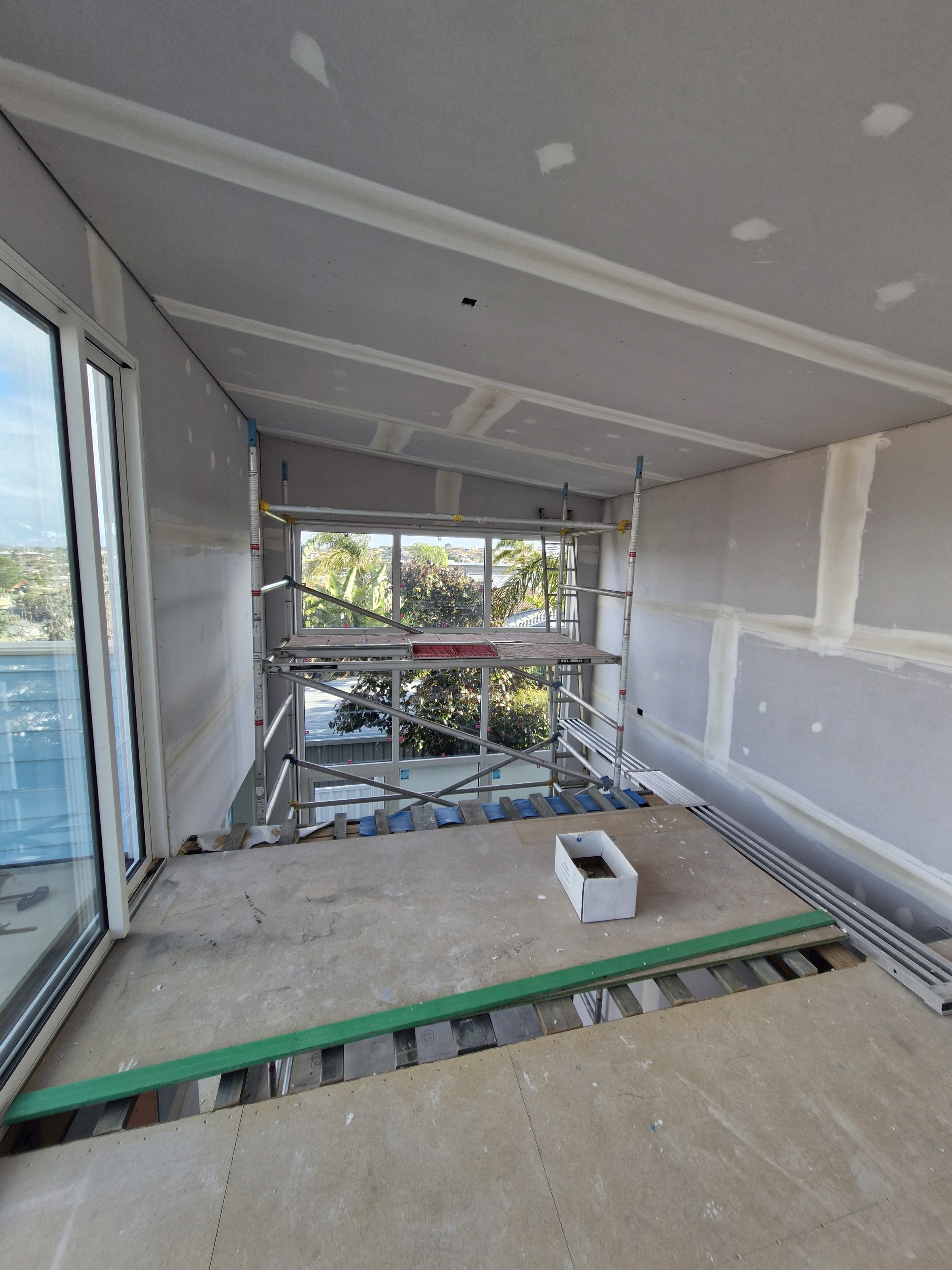 Interior of a building under construction with drywall walls and ceiling, scaffolding in front of large windows, and an unfinished floor with a piece of plywood and a small white box.