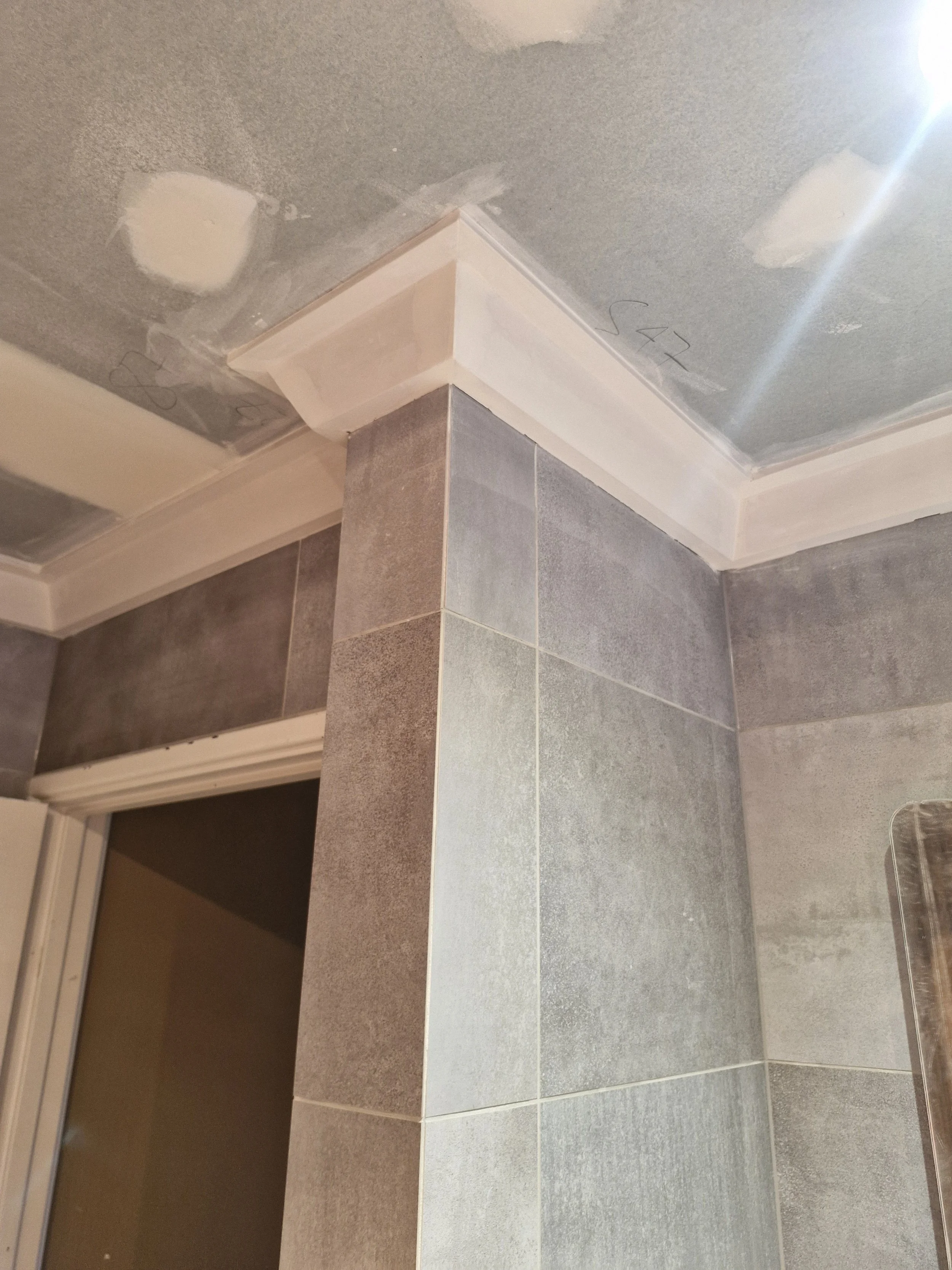 Close-up of a corner in a room under construction, showing tiled walls, drywall ceiling, and decorative molding with paint patches.