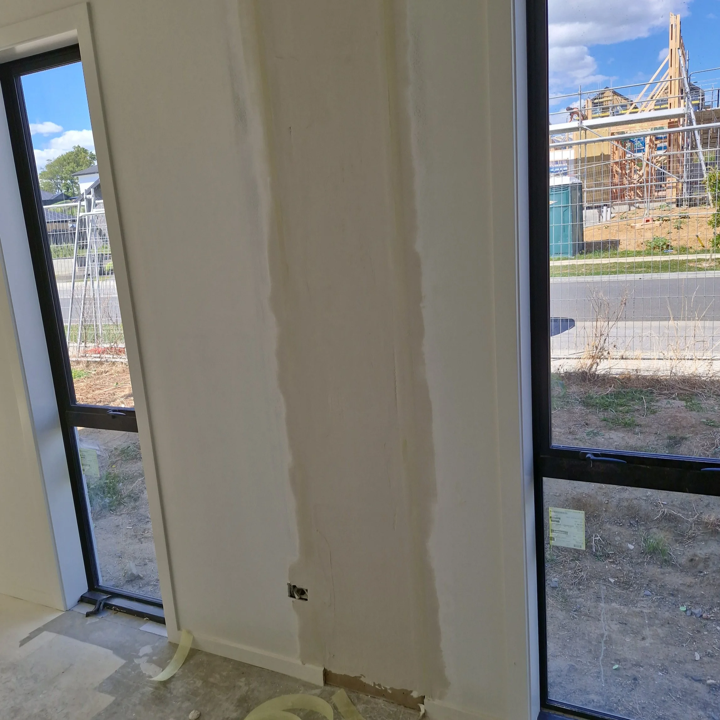 Interior wall with partially completed drywall installation and an electrical outlet box, next to large windows showing a construction site outside.