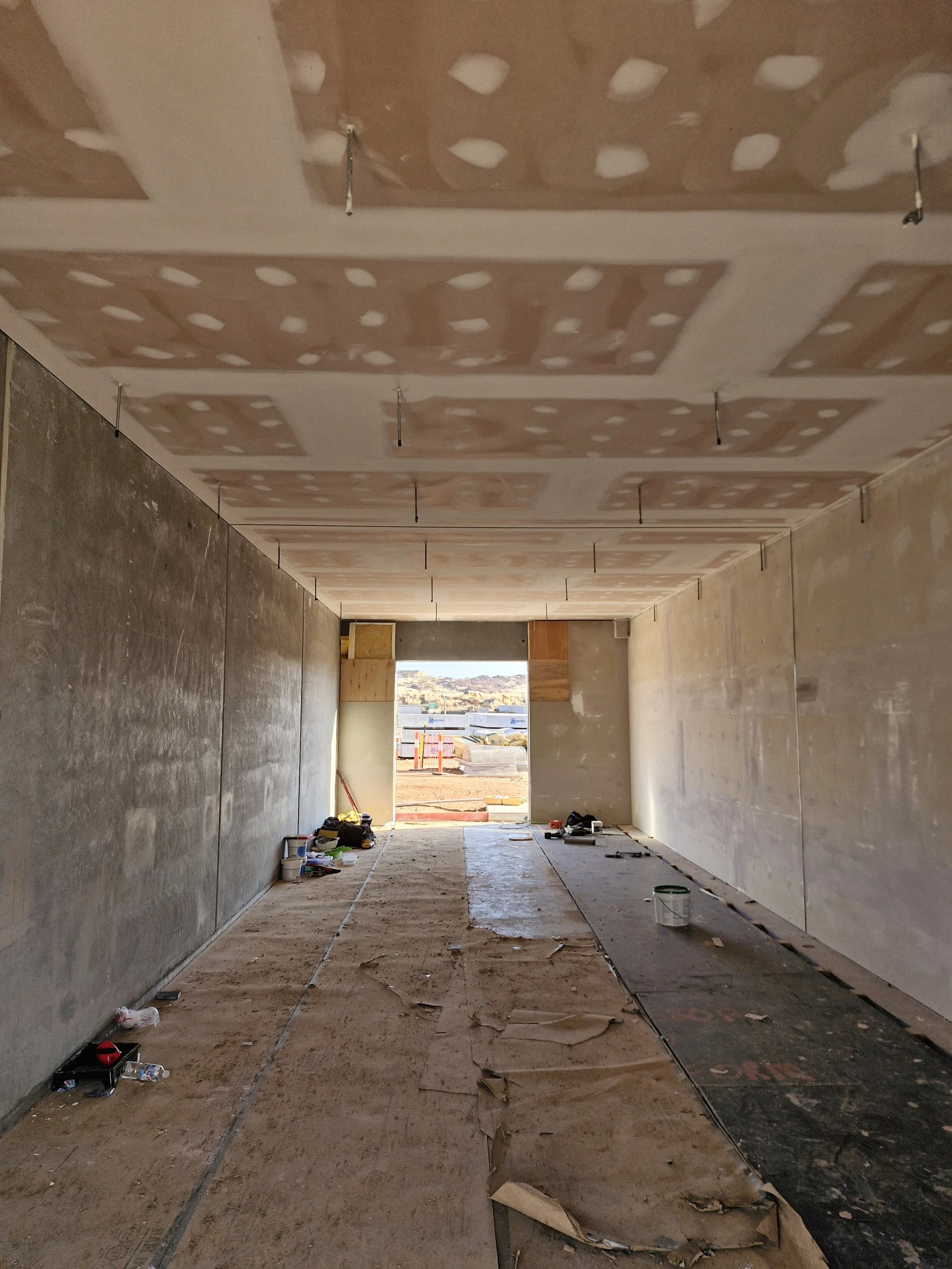 Interior of a house under construction with drywall ceiling and unfinished walls, construction tools and materials scattered on the floor, and a large opening leading outside to a construction site.