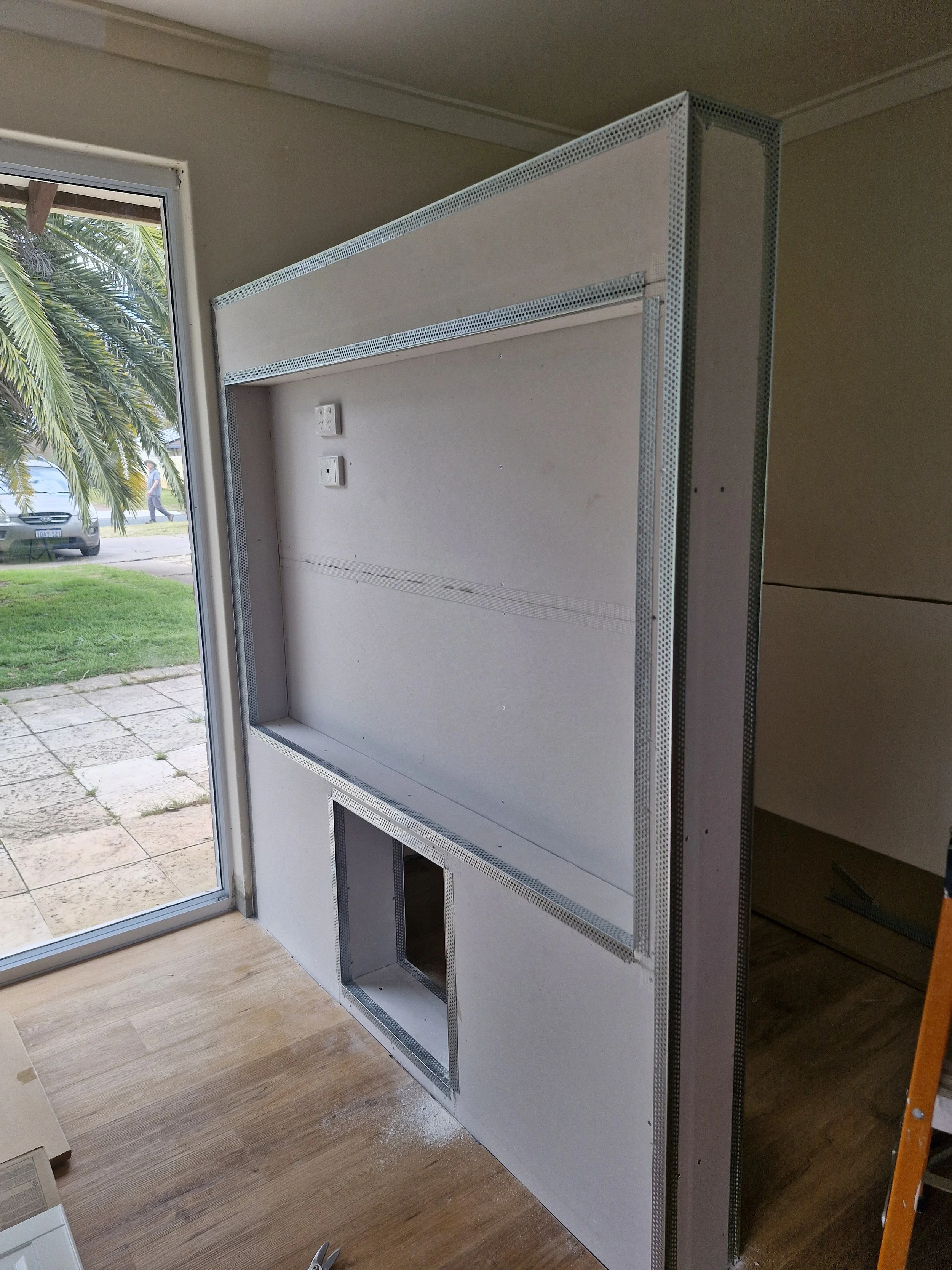 Interior structure of a wall with metal framing and electrical outlets, near a glass door opening to an outdoor patio with grass and a car visible outside.