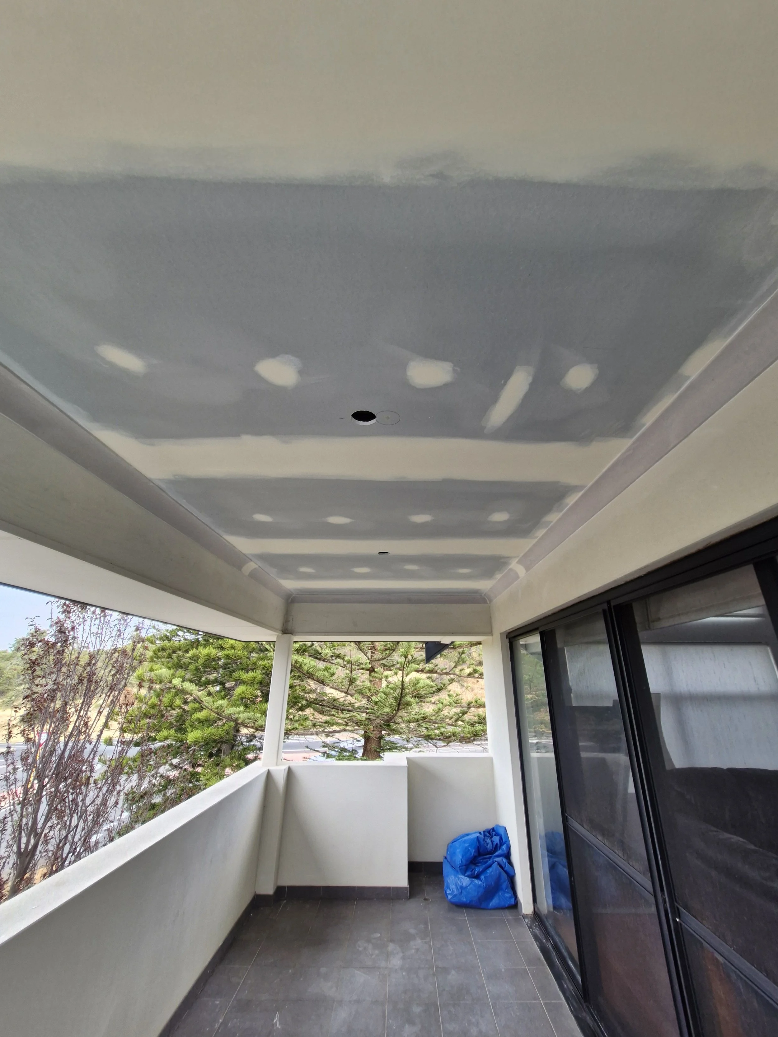 View of a balcony under renovation with drywall ceiling patching, a blue bag in the corner, and a sliding glass door leading to the apartment interior.