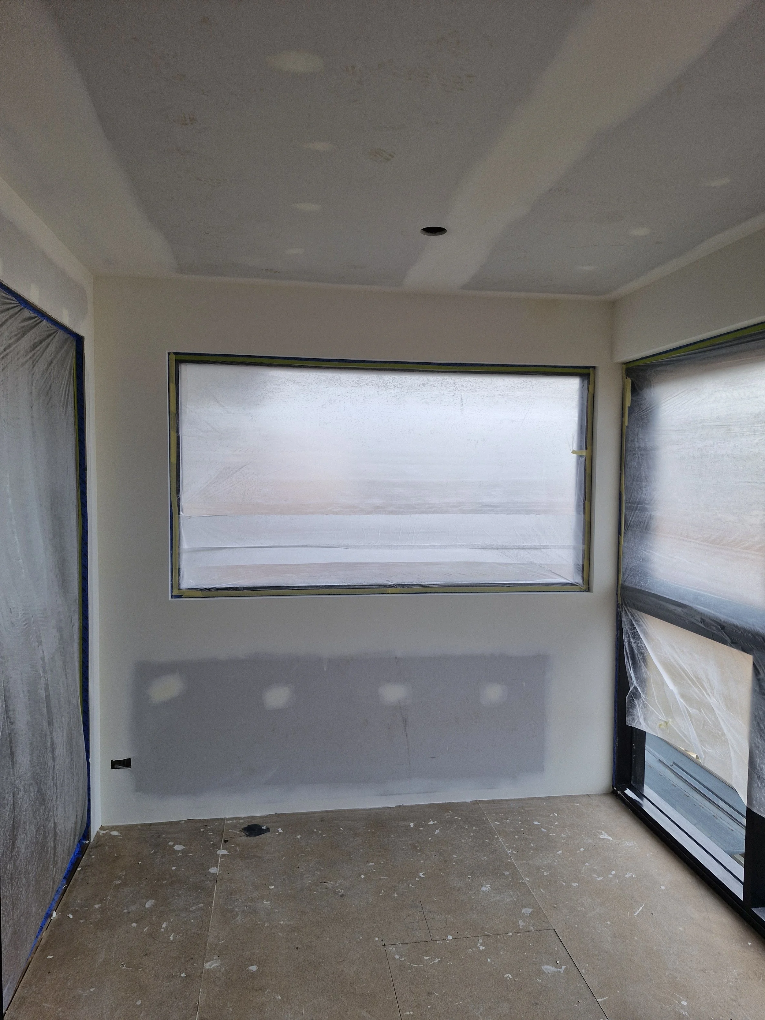An interior room under construction with unpainted drywall, taped and prepared for painting, with two large windows covered with protective plastic sheeting, and a ceiling with patches for lighting fixtures.