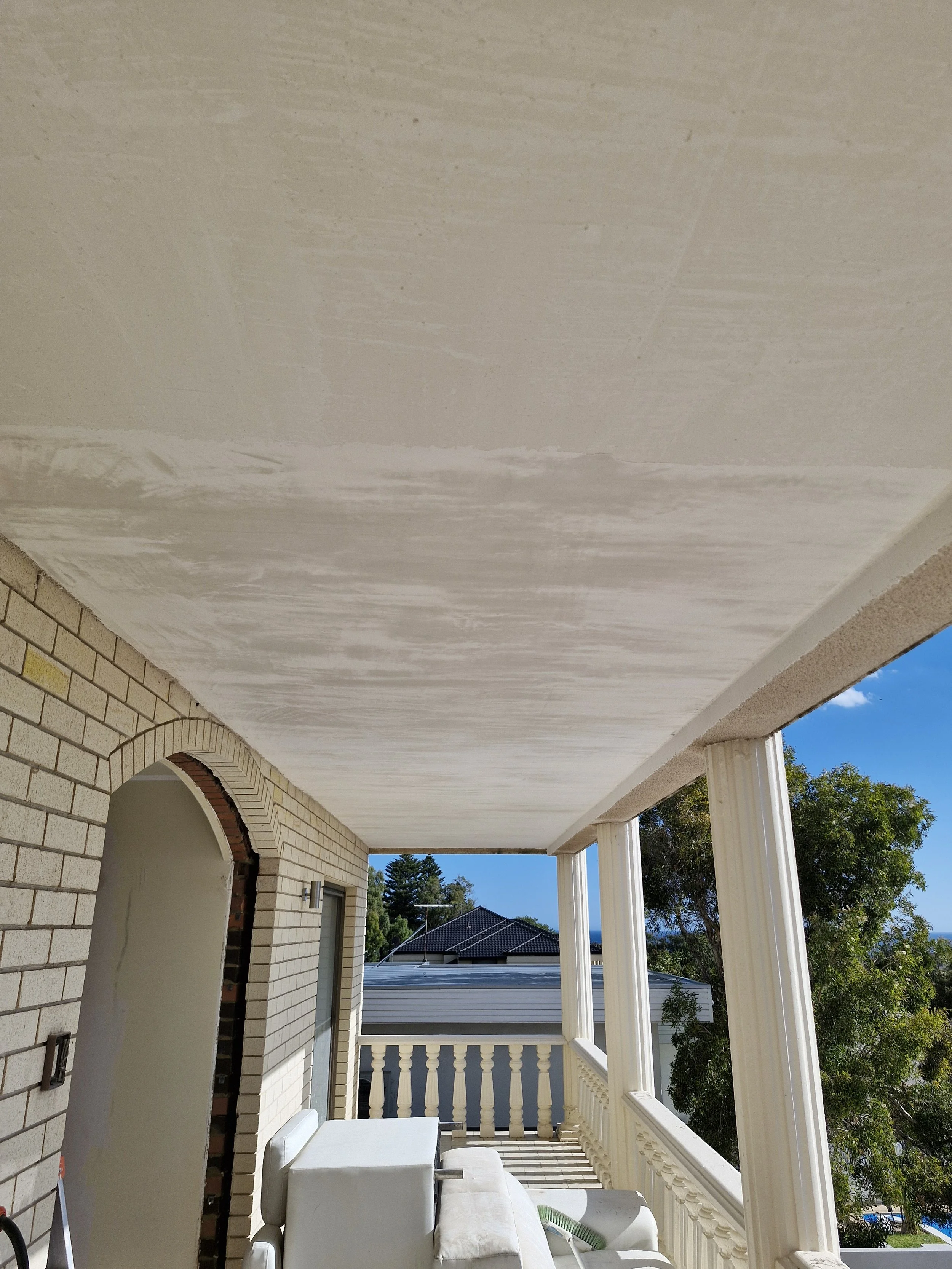 Covered porch with white railing and columns, outdoor furniture, brick wall, and view of trees and neighboring rooftops on a sunny day.