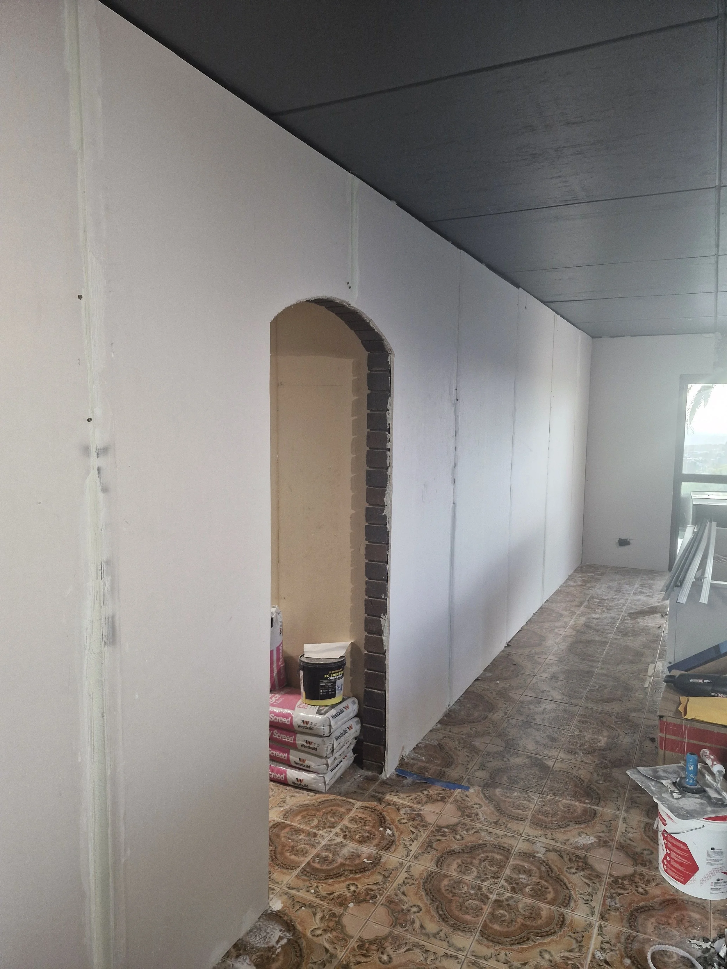 Interior under construction with partially finished walls, a small arched opening revealing building materials, and a window providing natural light.