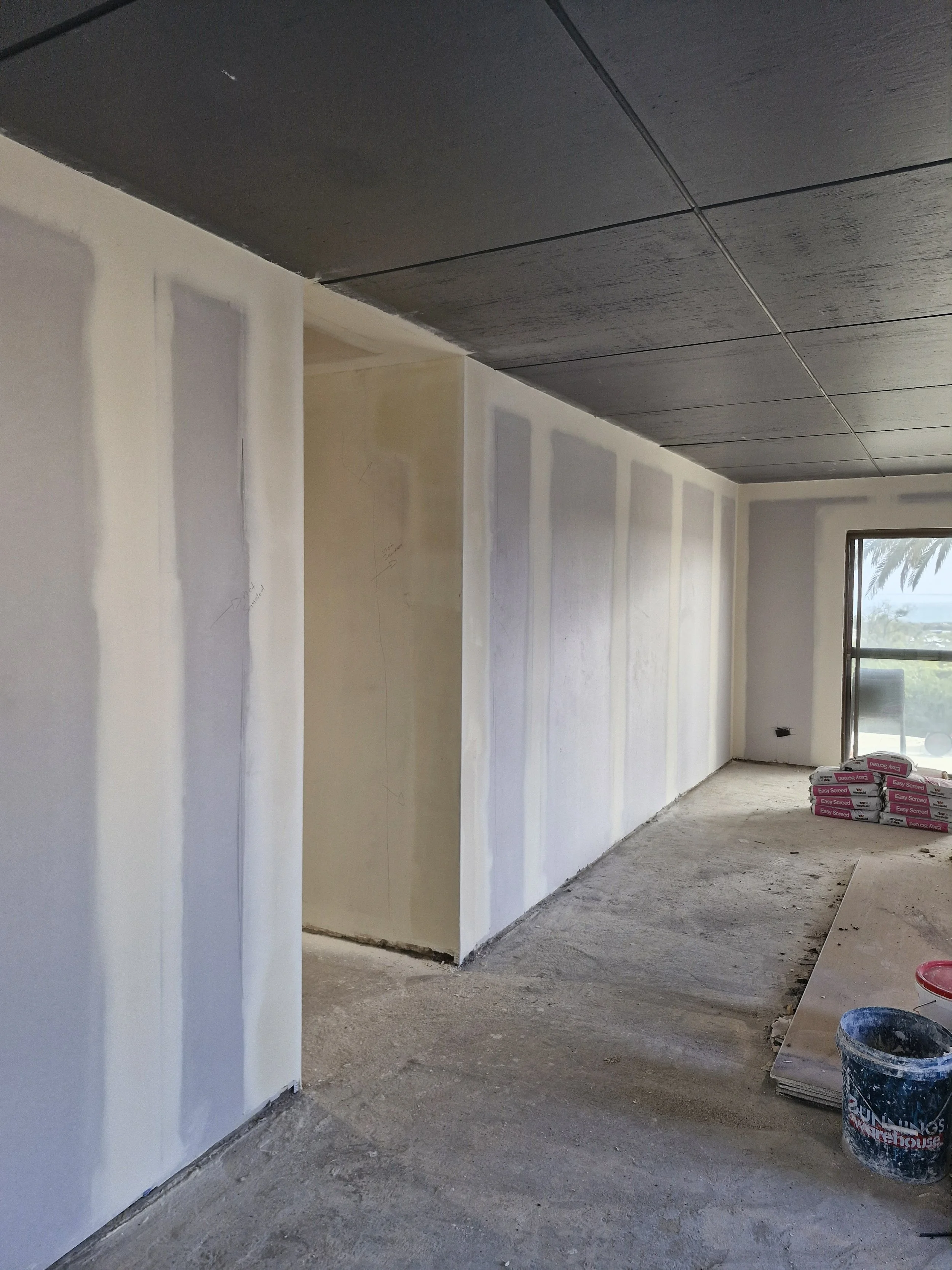 The interior of a room under construction with drywall partially installed and joint compound applied, construction supplies on the floor, and a window with a view outside.