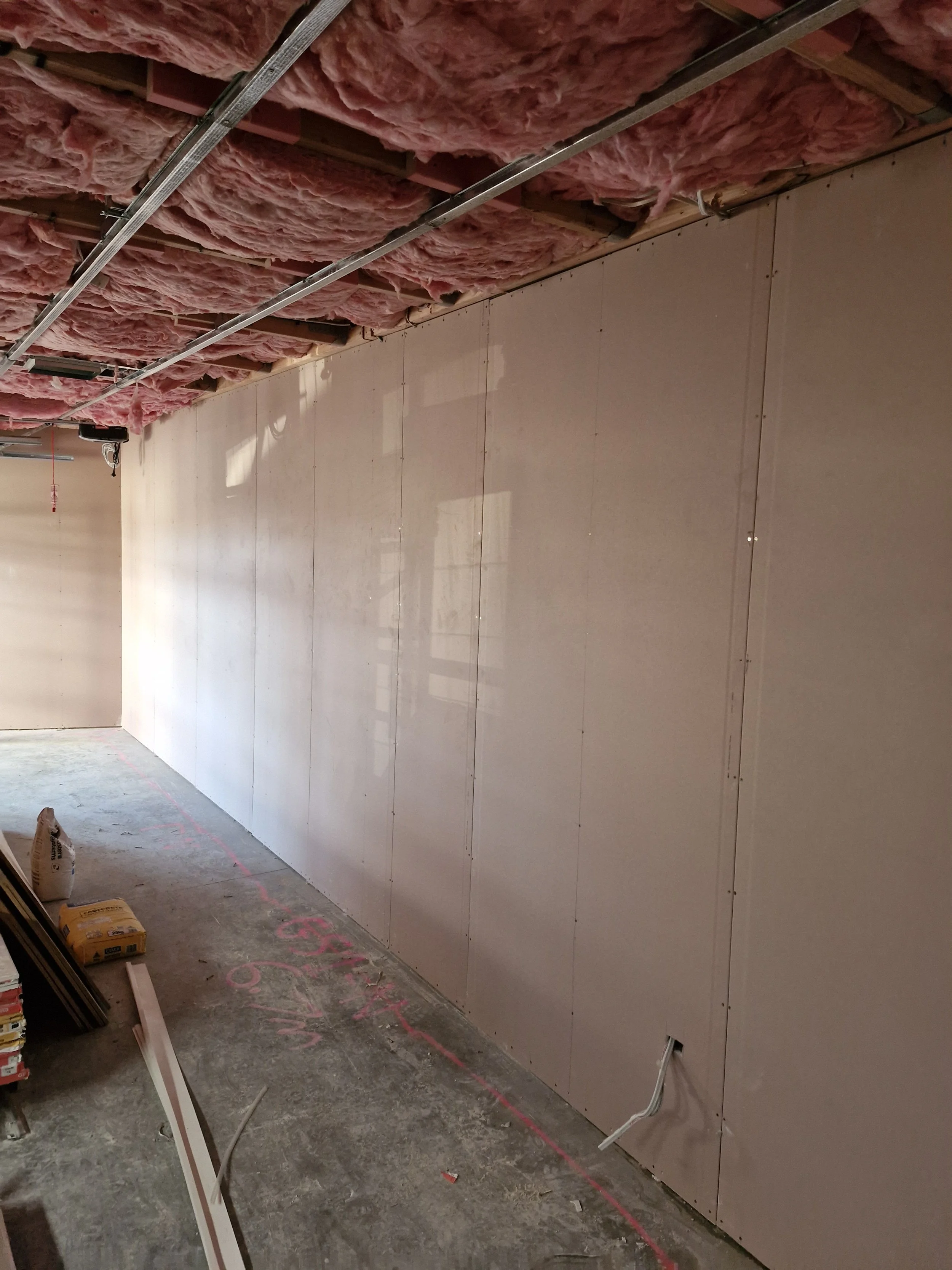 Construction site with drywall partially installed on a wall, pink insulation foam in the ceiling, and construction materials on the floor.