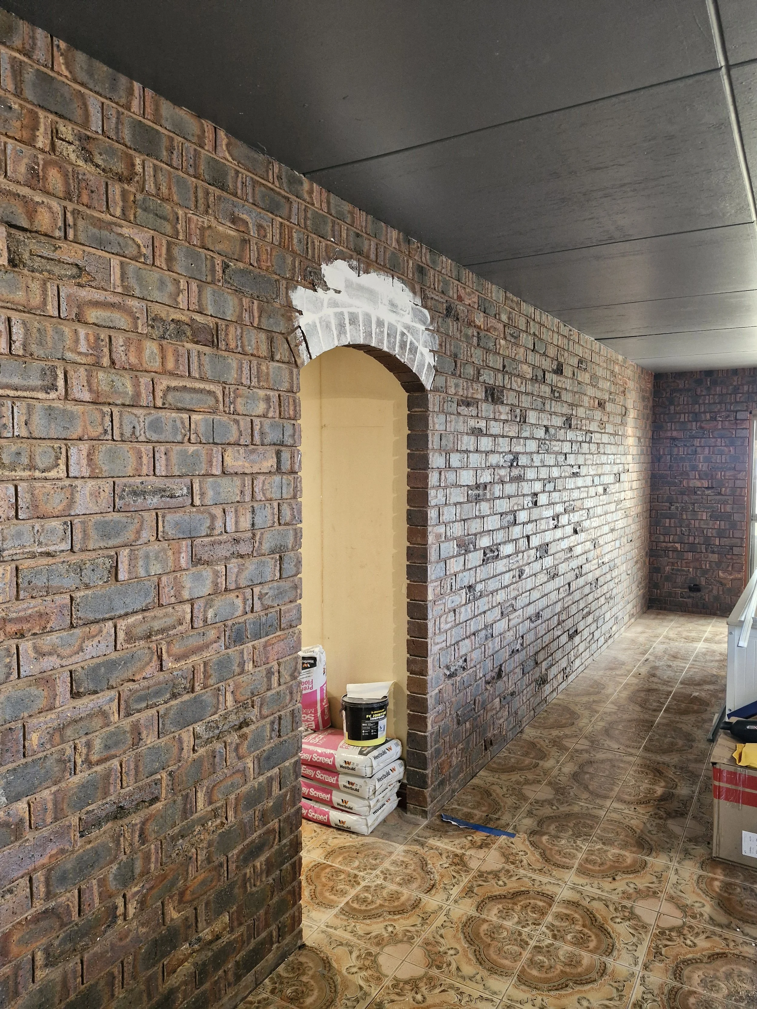 Interior of a room under renovation with exposed brick walls, some painted white, and patterned tile flooring. Construction materials are stacked near an arched doorway.