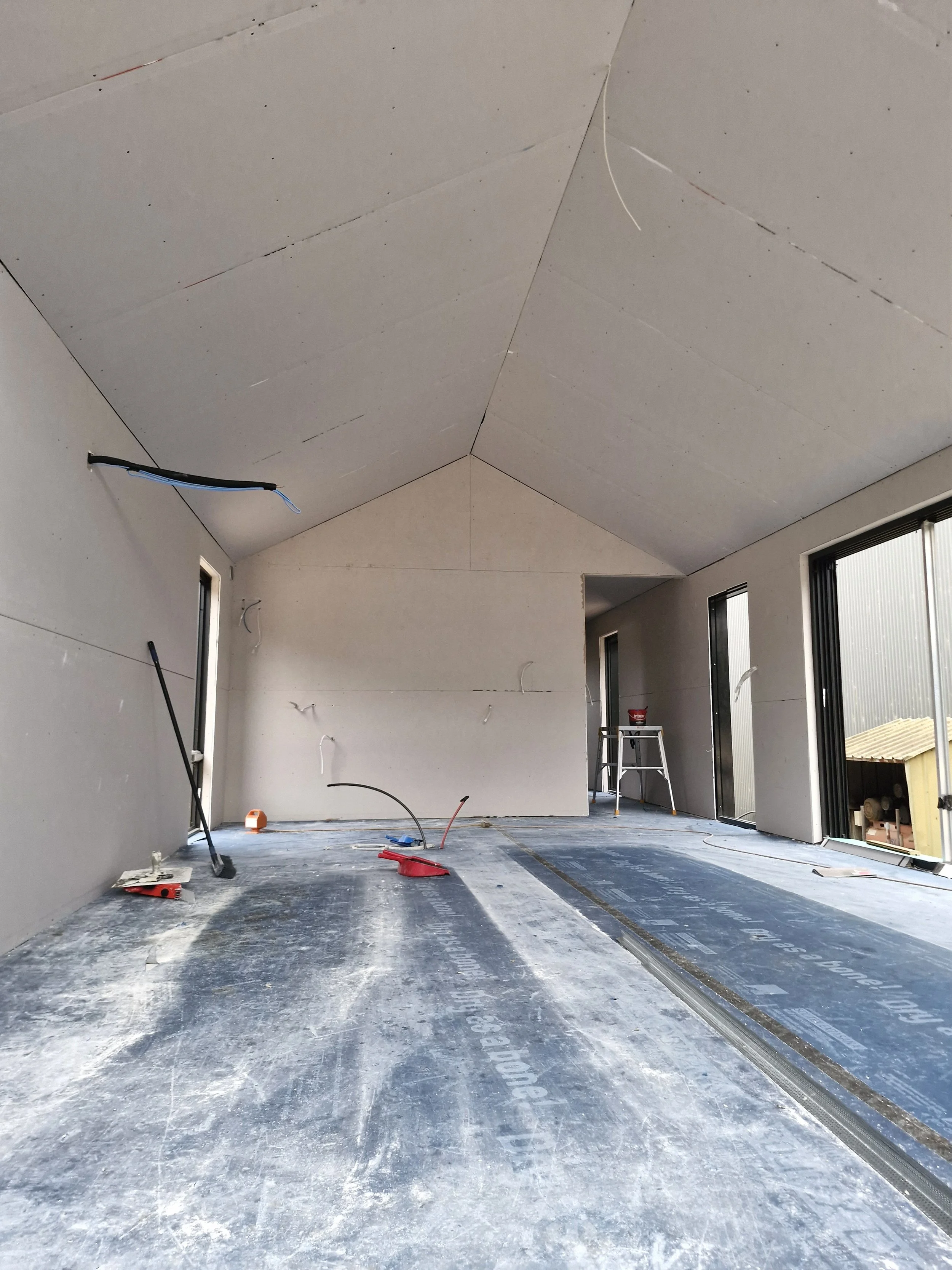 Interior of a room under construction with drywall panels on the walls and ceiling, some wiring visible, and construction tools scattered on the floor.