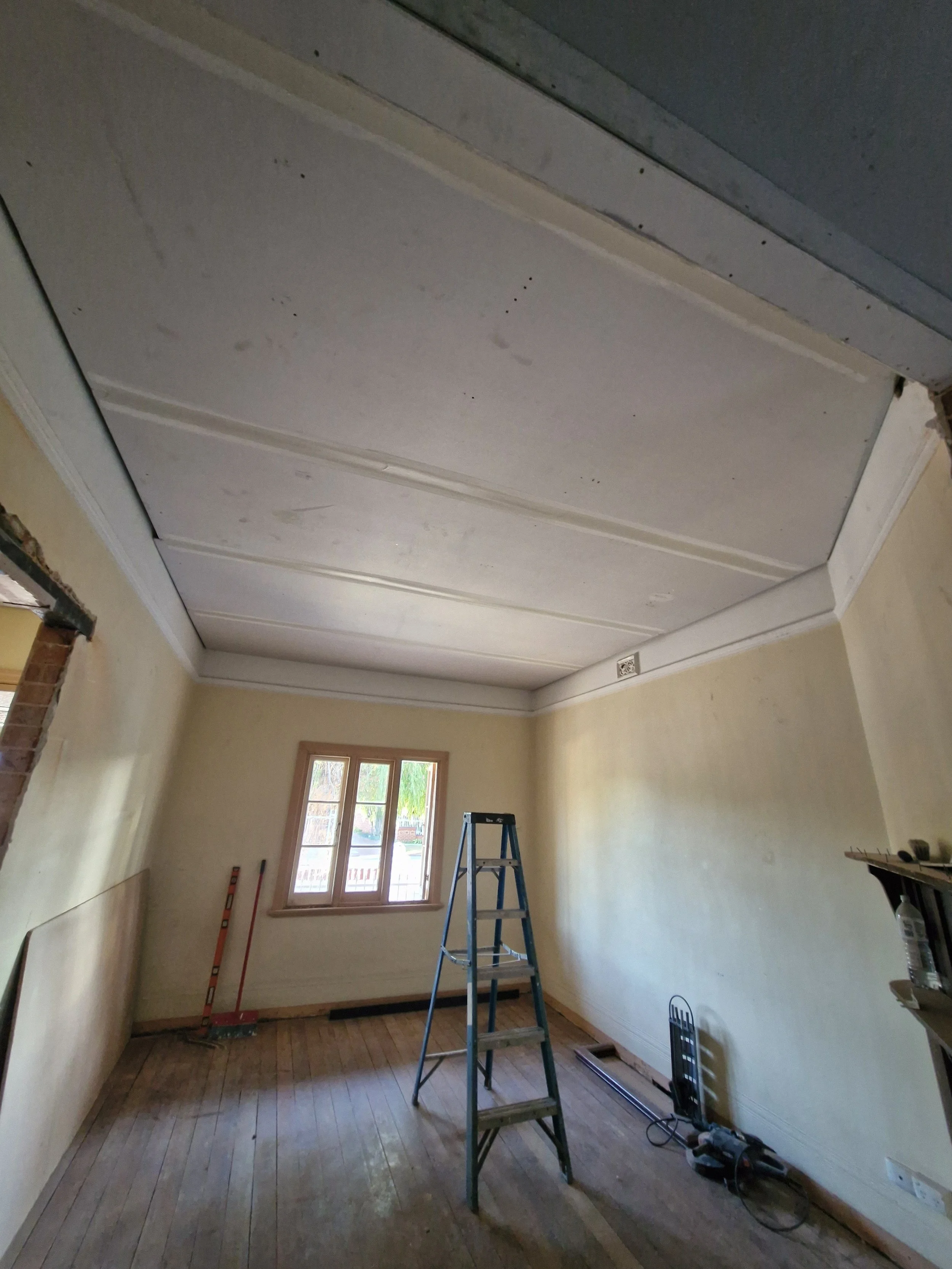 A room undergoing renovation with a ladder in the center, construction tools on the floor, and a window letting in natural light. The ceiling is partly unfinished with visible seams, and the walls are painted light yellow.