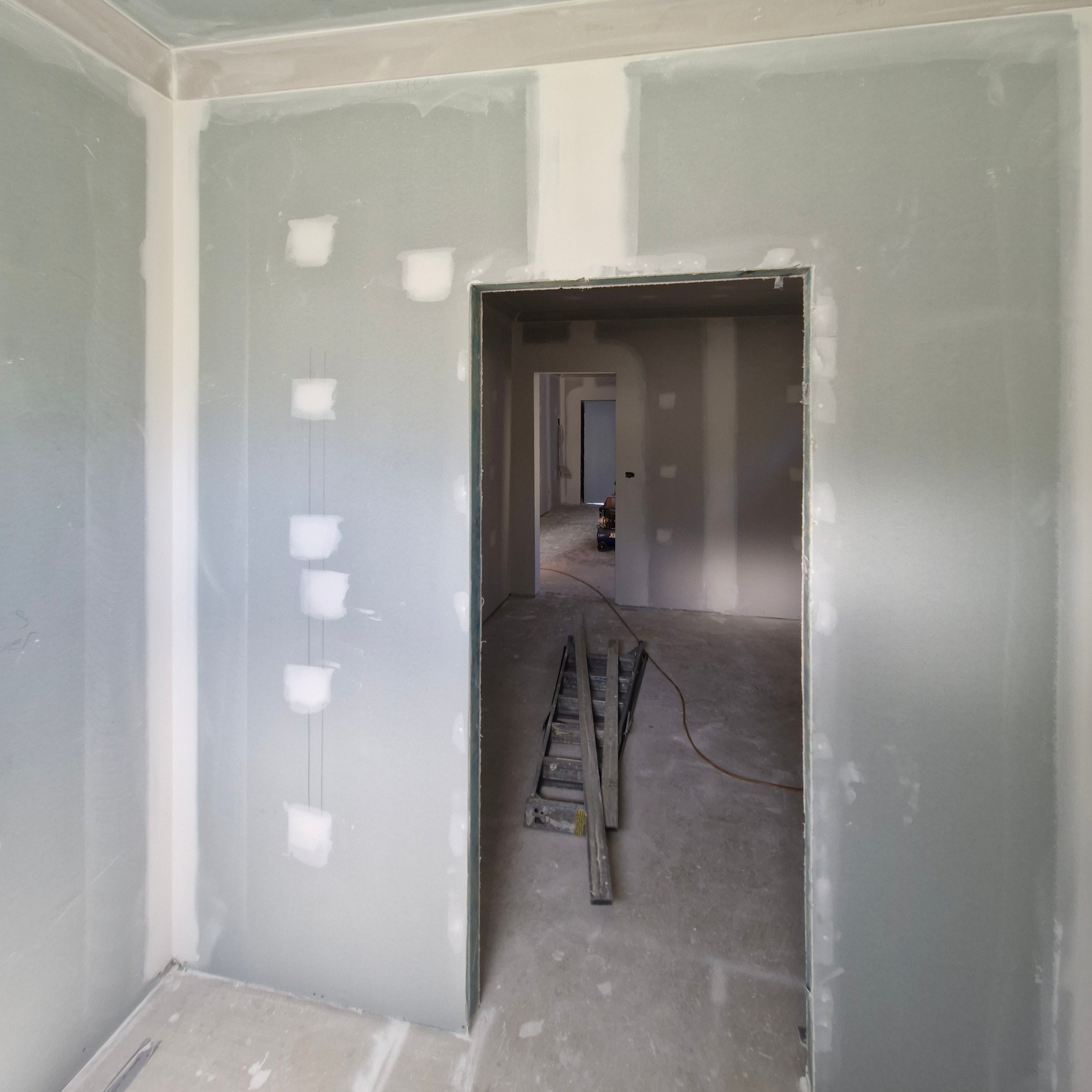 An unfinished room with drywall installation in progress. The walls are partially covered with drywall, and there is a doorway leading into another room. Construction tools and materials are present on the floor.