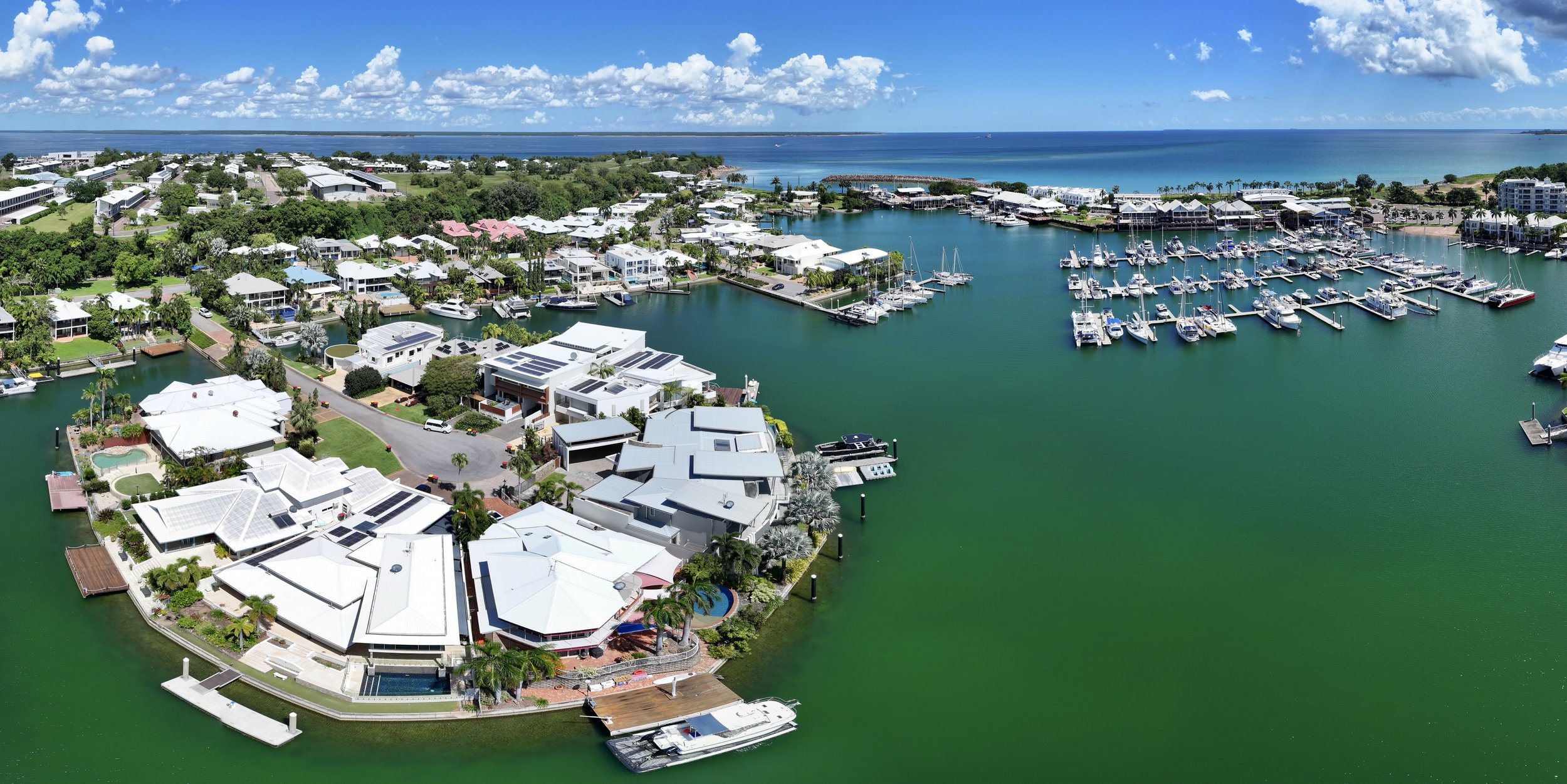 Aerial view of a marina with boats, waterfront houses with white roofs, and a distant ocean shoreline under a partly cloudy sky.