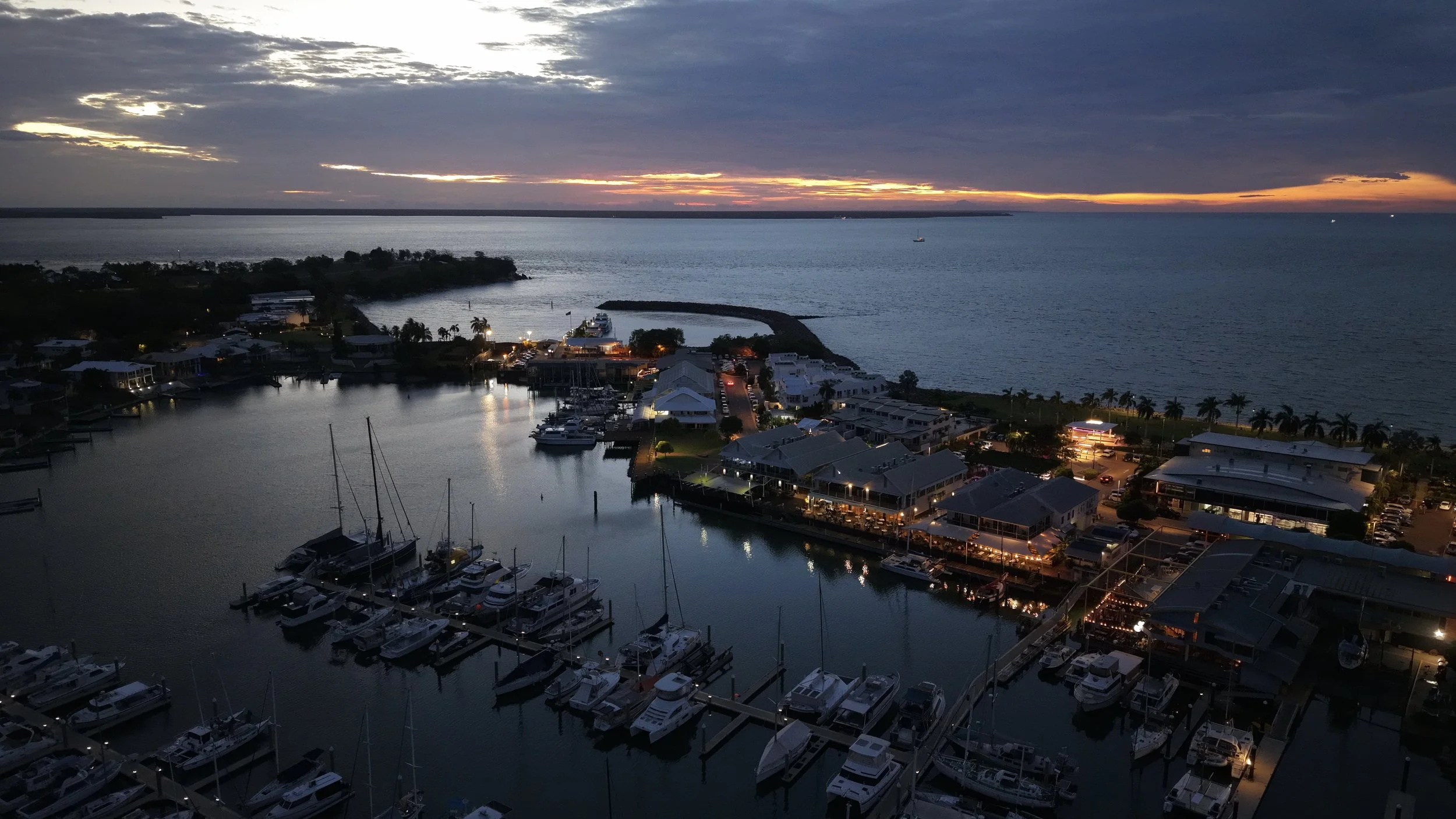 Aerial view of a marina at sunset with boats docked, buildings along the shore, palm trees, and the ocean in the background, under a partly cloudy sky.