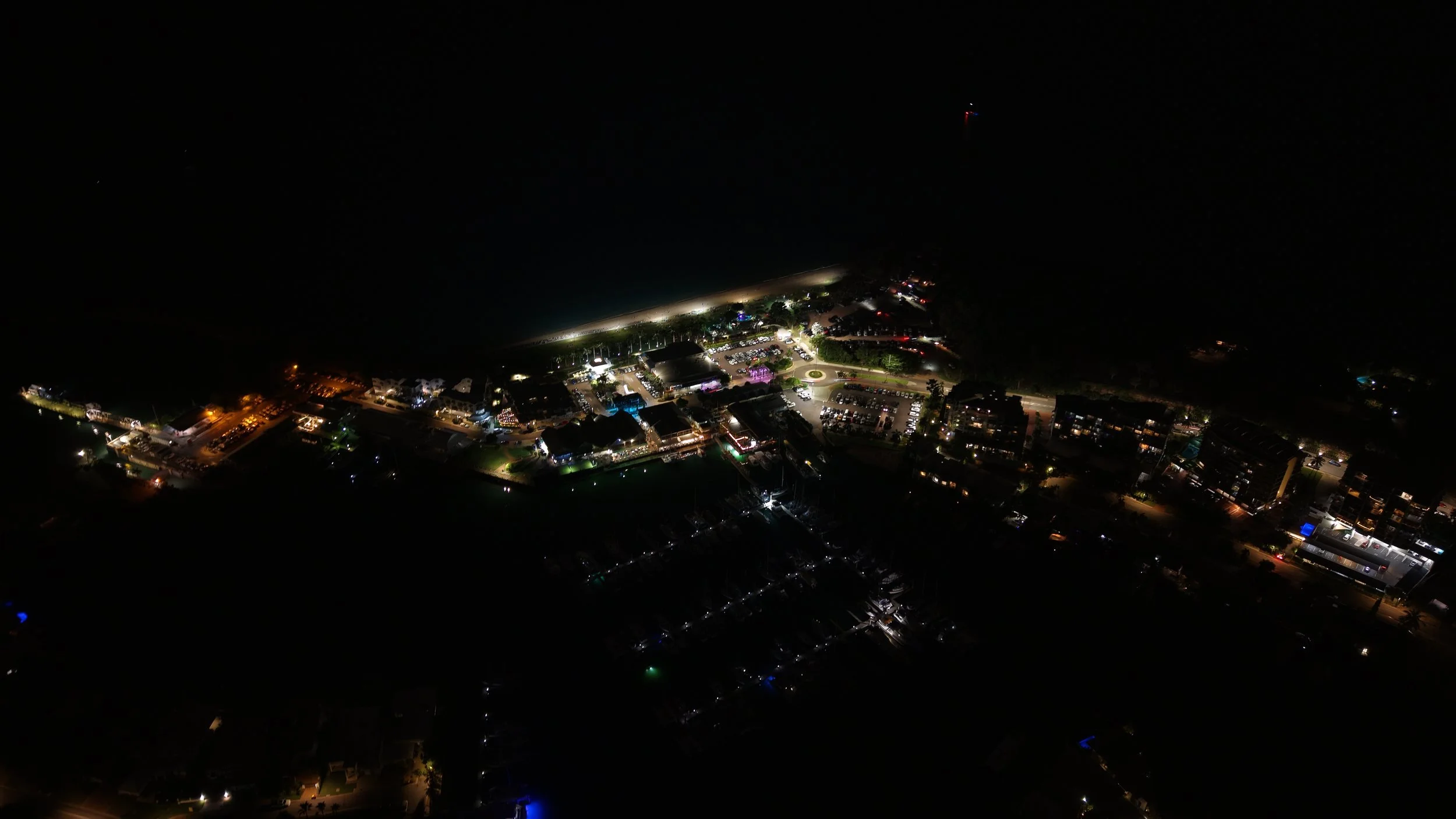Night aerial view of a brightly lit coastal area with buildings, parking lots, and streetlights.