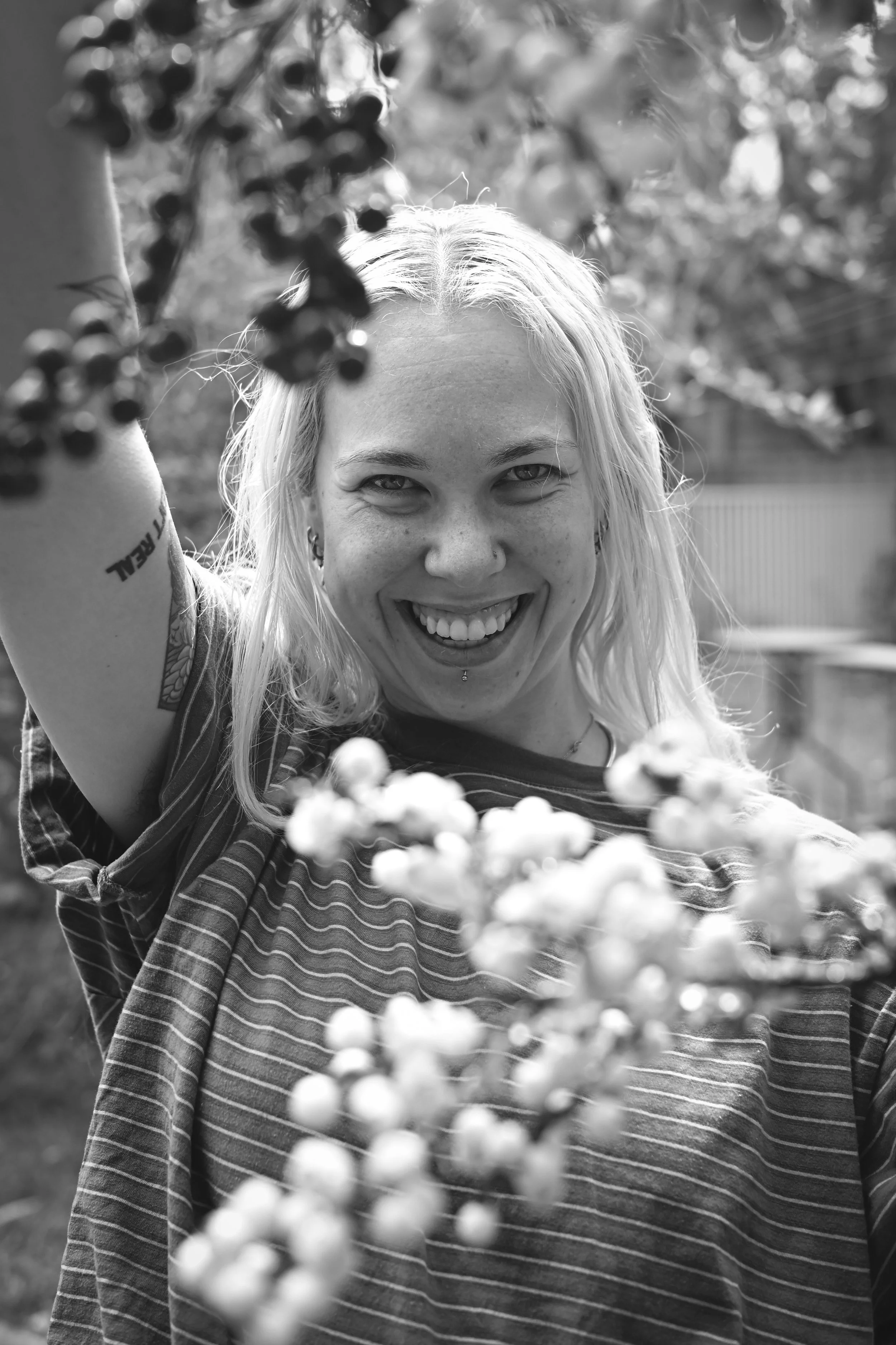 A smiling woman with long light-colored hair, wearing a striped shirt, standing outdoors among trees and berries, with a blurred background.