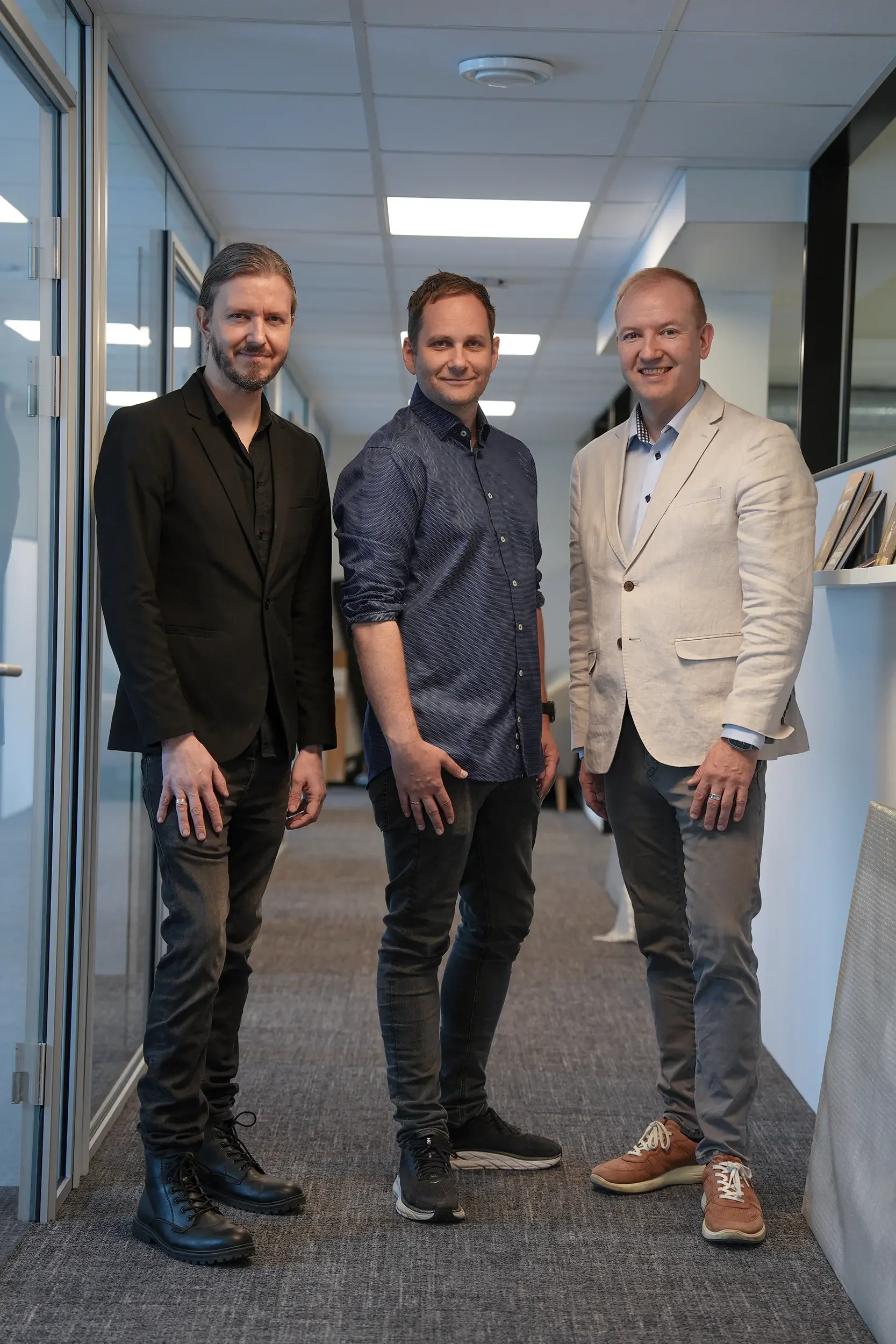 Tre menn står i en kontorgang, smilende og iført forretningskledd, hver med ulike sko.