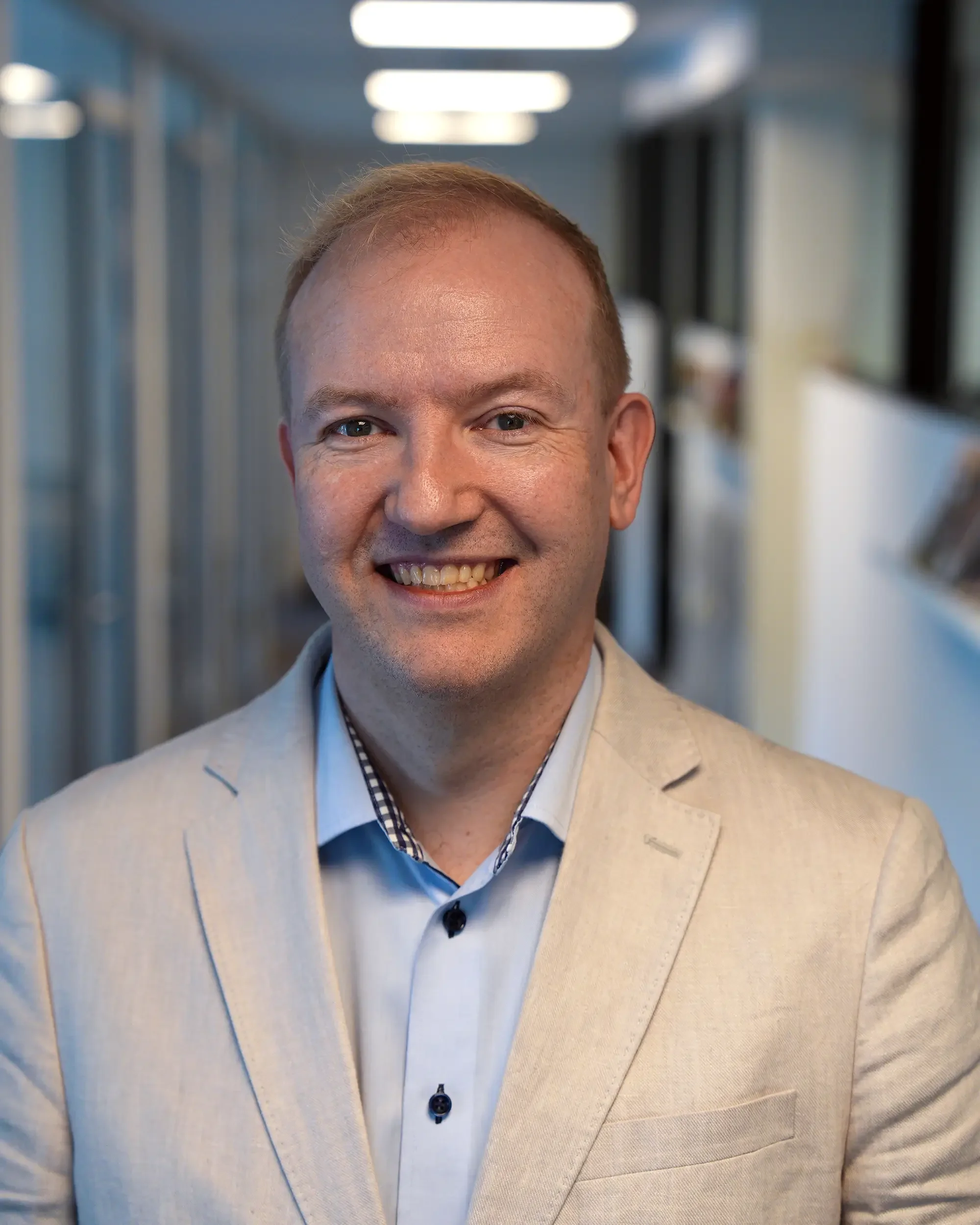 Nærbilde av en mann i en lys blazer og blå skjorte, smilende, i en korridor med taklys. Geir Arne Solheim fra Mediehub Group AS.