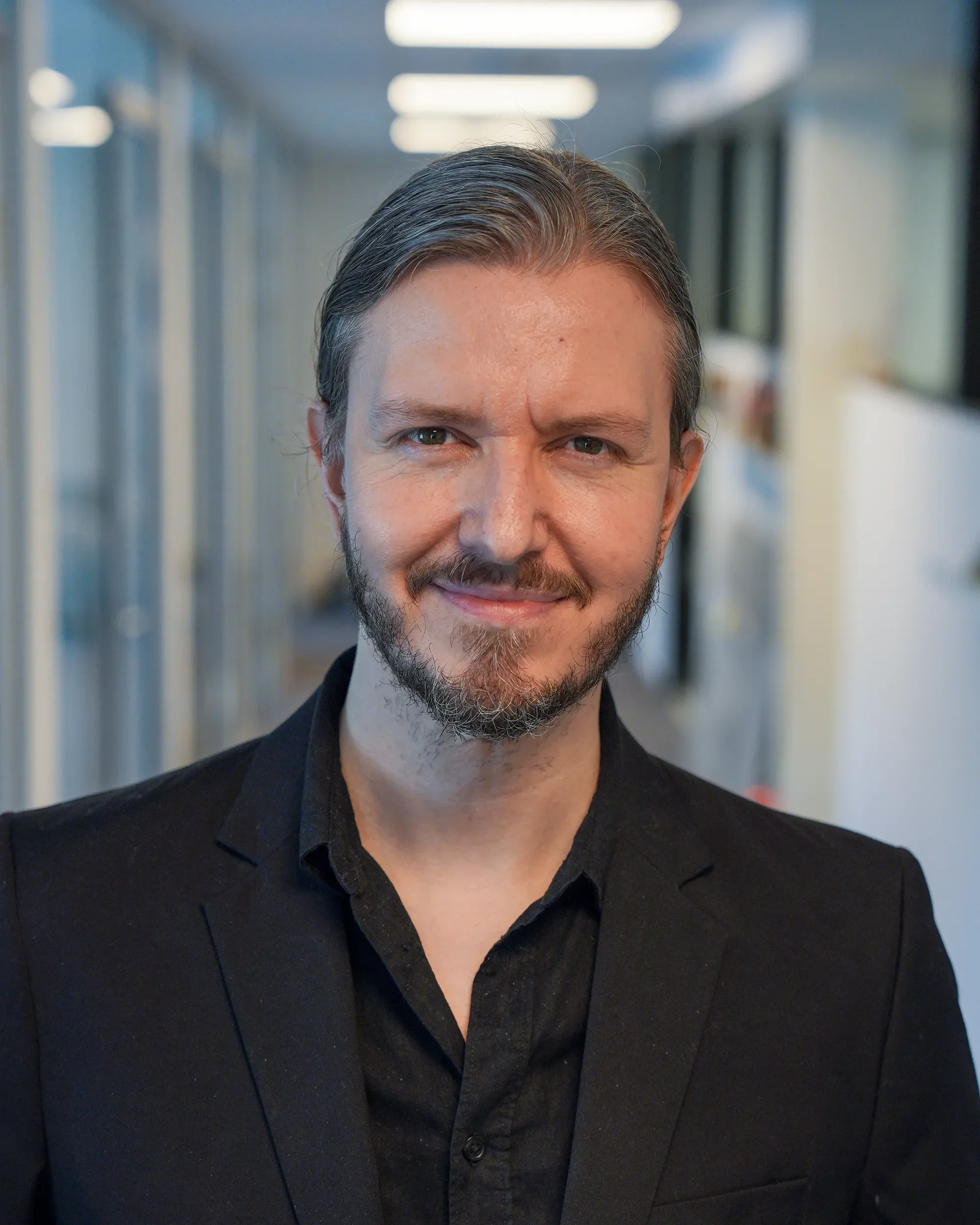 Et nærbilde av en mann med brunt og grått hår og skjegg, iført svart dress og skjorte, som smiler i en innendørs korridor.
