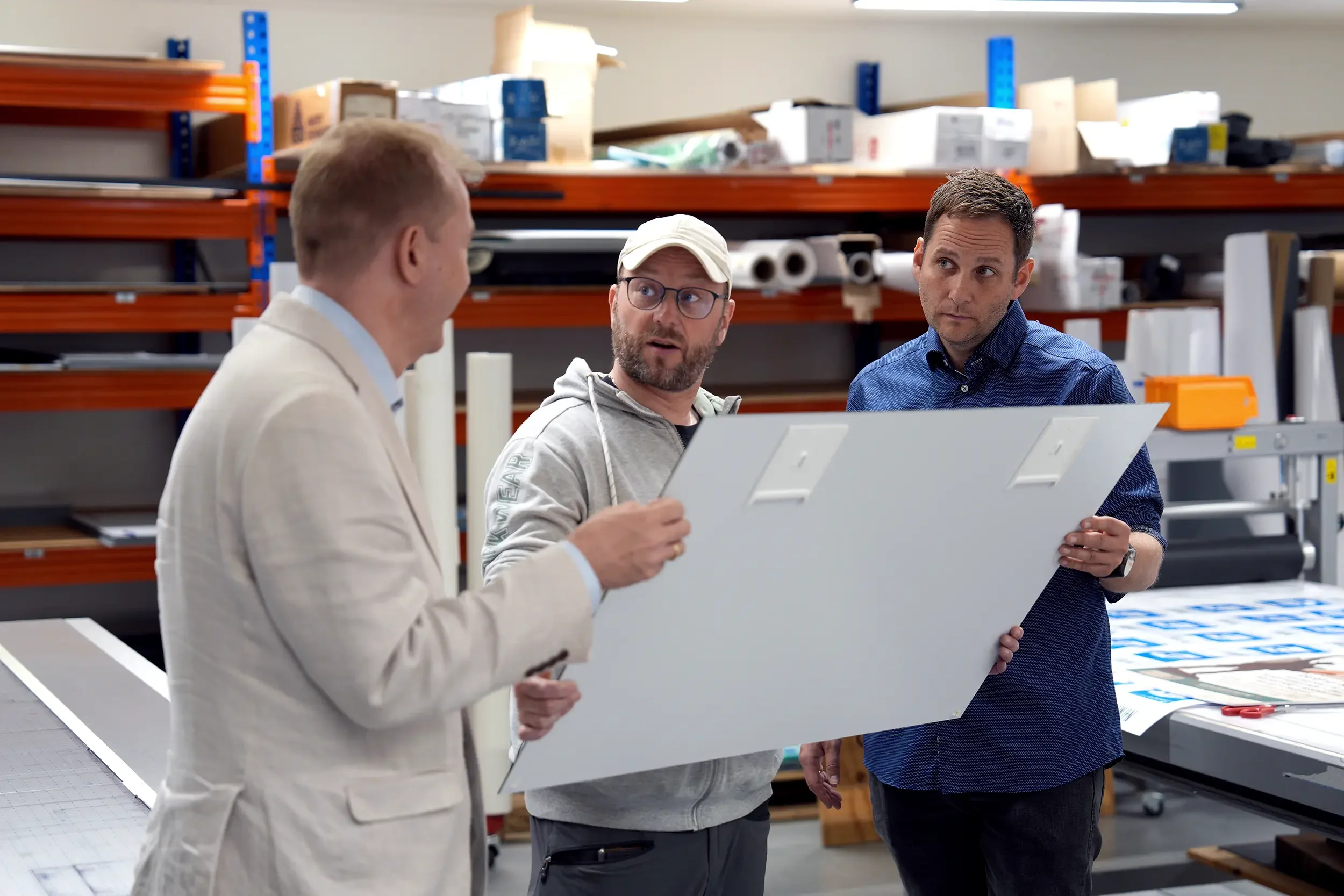 Tre menn i industriell setting som diskuterer et stort hvitt panel.